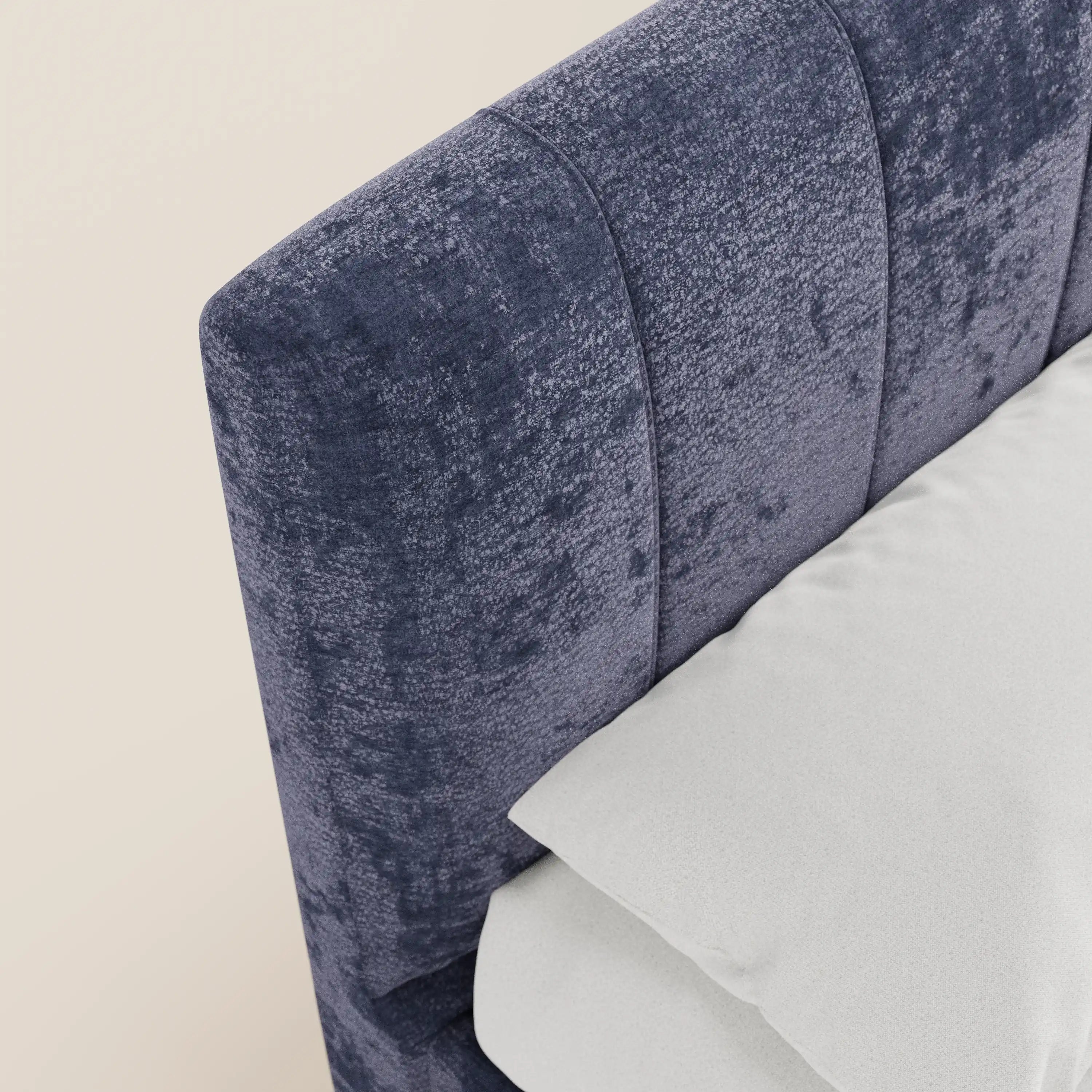 Un primo piano del letto imbottito ONYX di Letti.store mette in risalto la testiera in tessuto blu scuro, certificata Oeko-Tex®, con cuciture verticali, che si abbina perfettamente alla biancheria da letto bianca su uno sfondo bianco sporco.