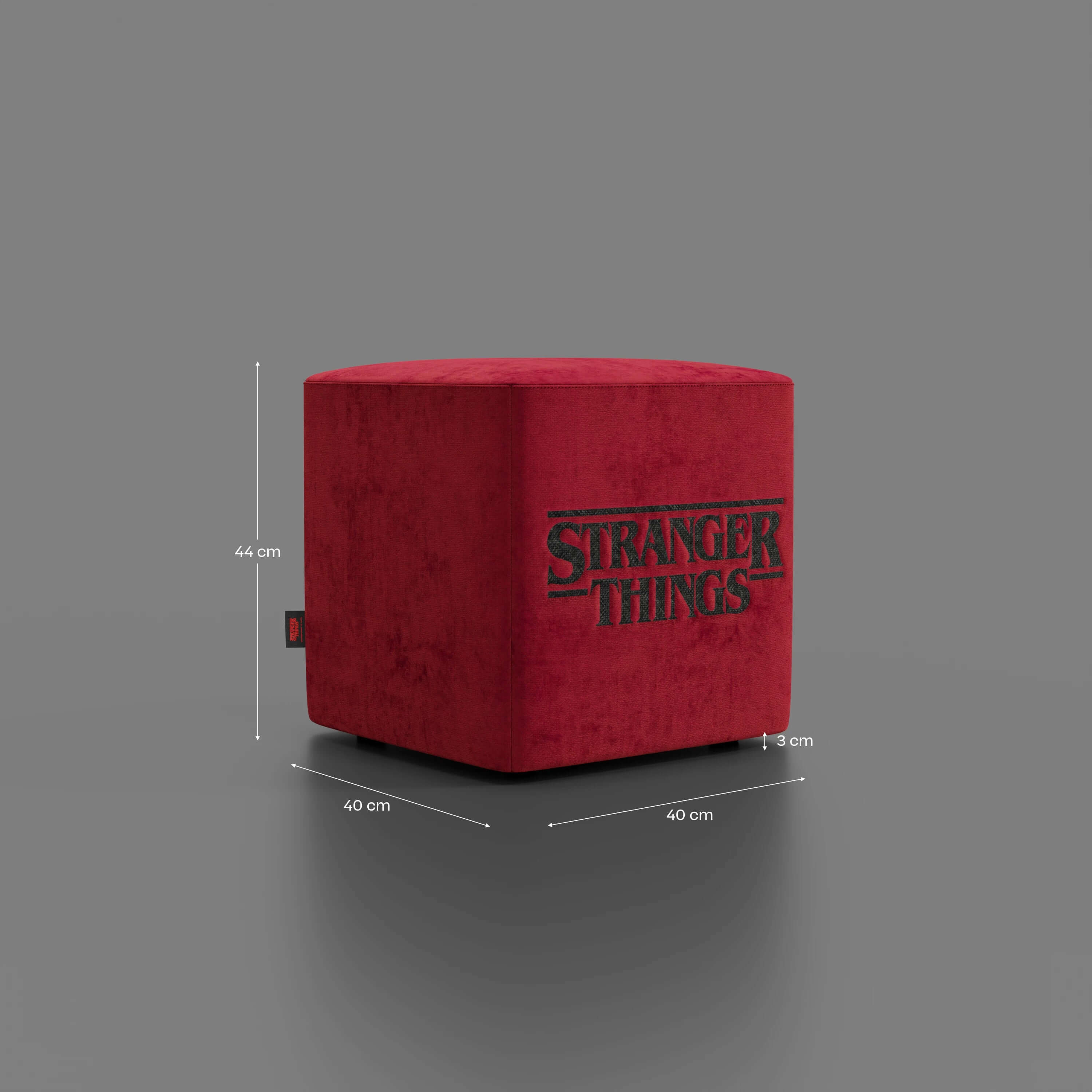 Il pouf Cube Stranger Things Edition di Divani.Store è di colore rosso, con il logo della serie e una piccola etichetta nera, su sfondo grigio. Realizzato in tessuto antimacchia, misura 44x40x40 cm.