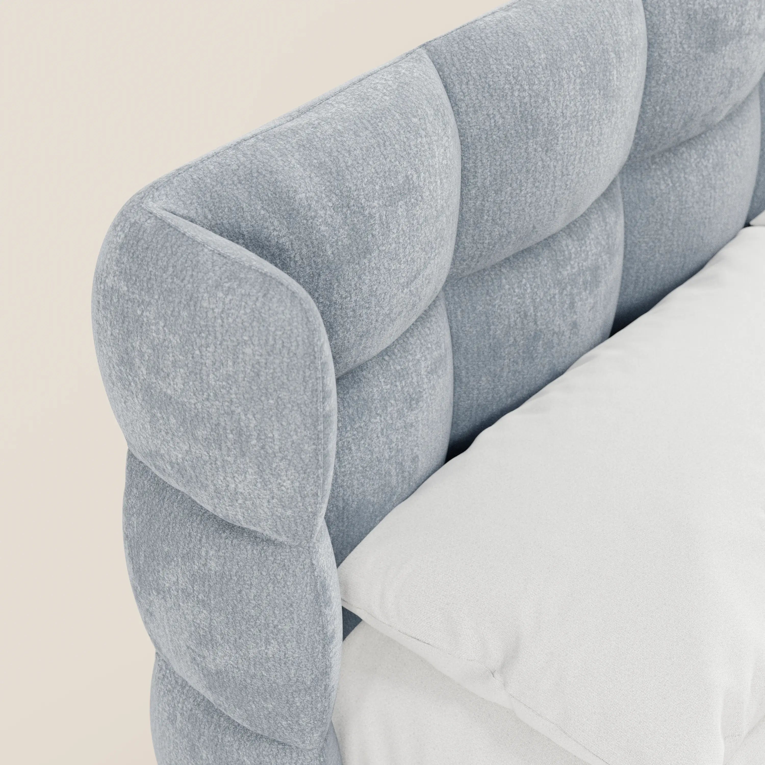 Primo piano del letto imbottito RAGNAR di Letti.store, caratterizzato da una testiera avvolgente capitonné azzurra, realizzata in un elegante tessuto certificato Oeko-Tex®. Sul letto sono appoggiati un cuscino bianco e un lenzuolo coordinato; lo sfondo è di un beige chiaro.