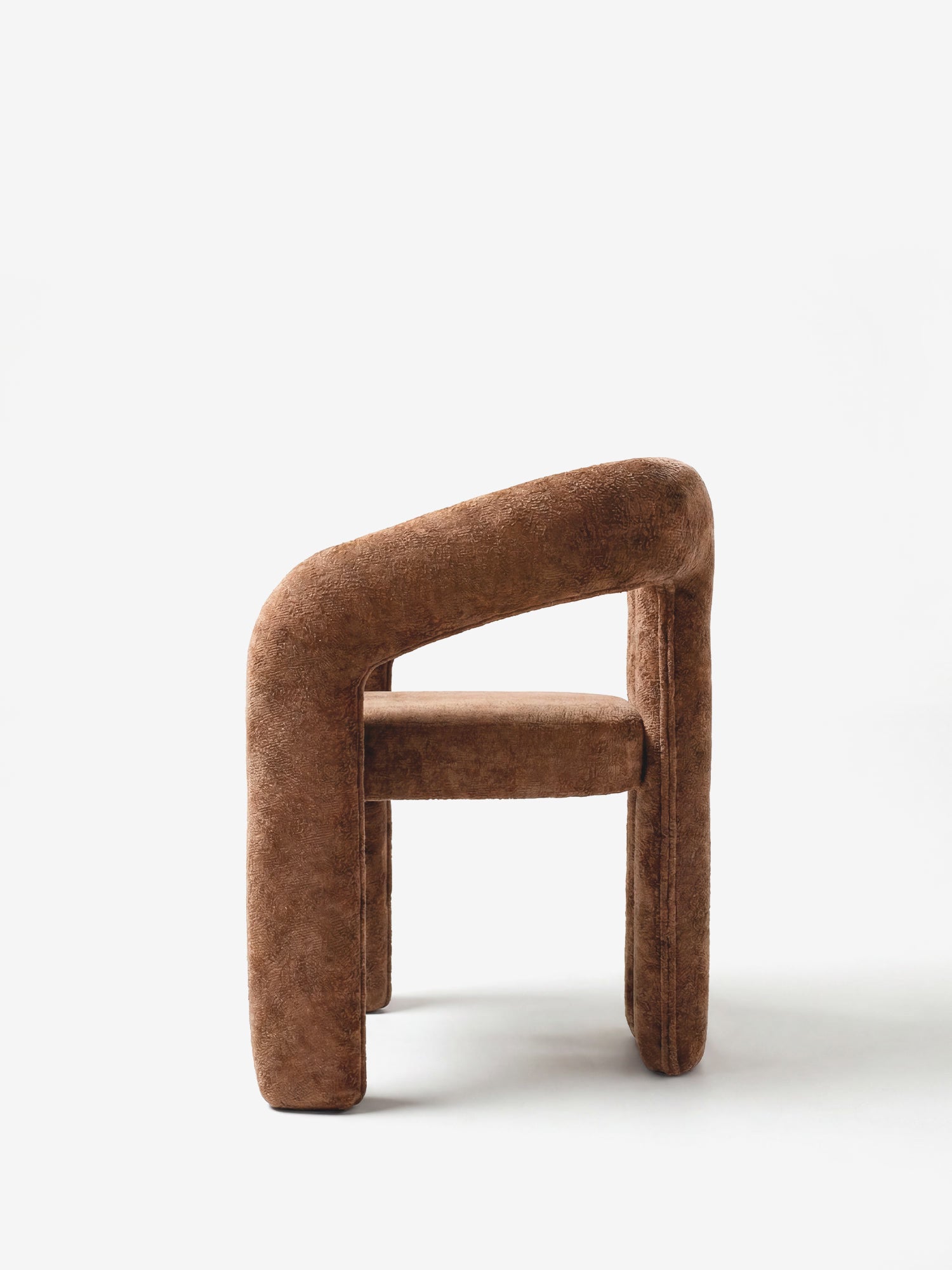La sedia di design imbottita Sibilla di Divani.Store è caratterizzata da un morbido tessuto marrone, uno schienale curvo e braccioli che formano un arco, oltre a gambe e seduta spesse e arrotondate, che le conferiscono un aspetto audace e scultoreo su uno sfondo bianco a tinta unita.