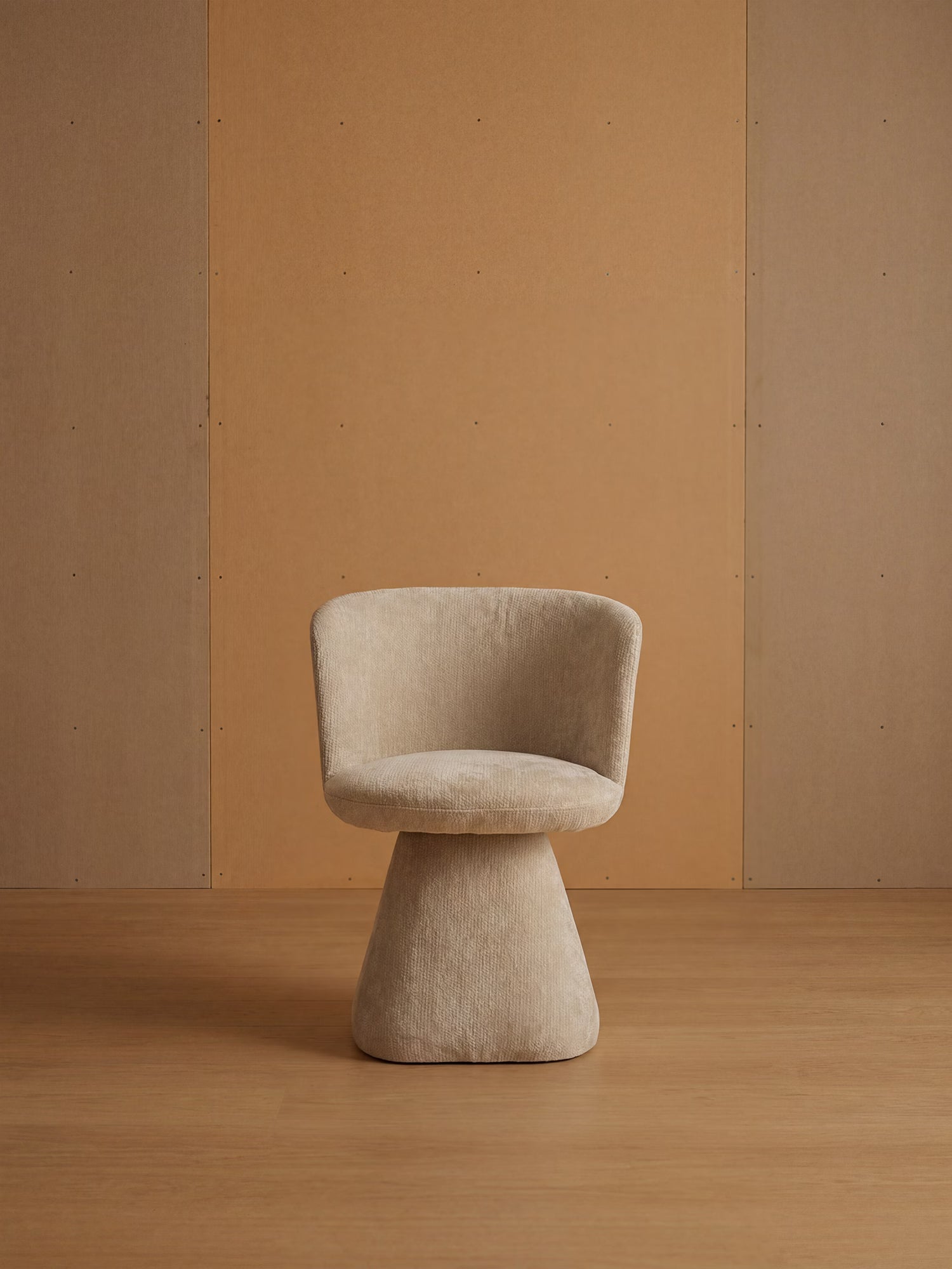 La sedia moderna Talia, imbottita in morbido tessuto ciniglia OEKO-TEX® di Divani.Store, caratterizzata da uno schienale ricurvo e da una base imbottita, è posizionata su un pavimento in legno chiaro tra pannelli color cuoio: una scena minimalista dall’eleganza sobria e dai toni caldi e neutri.