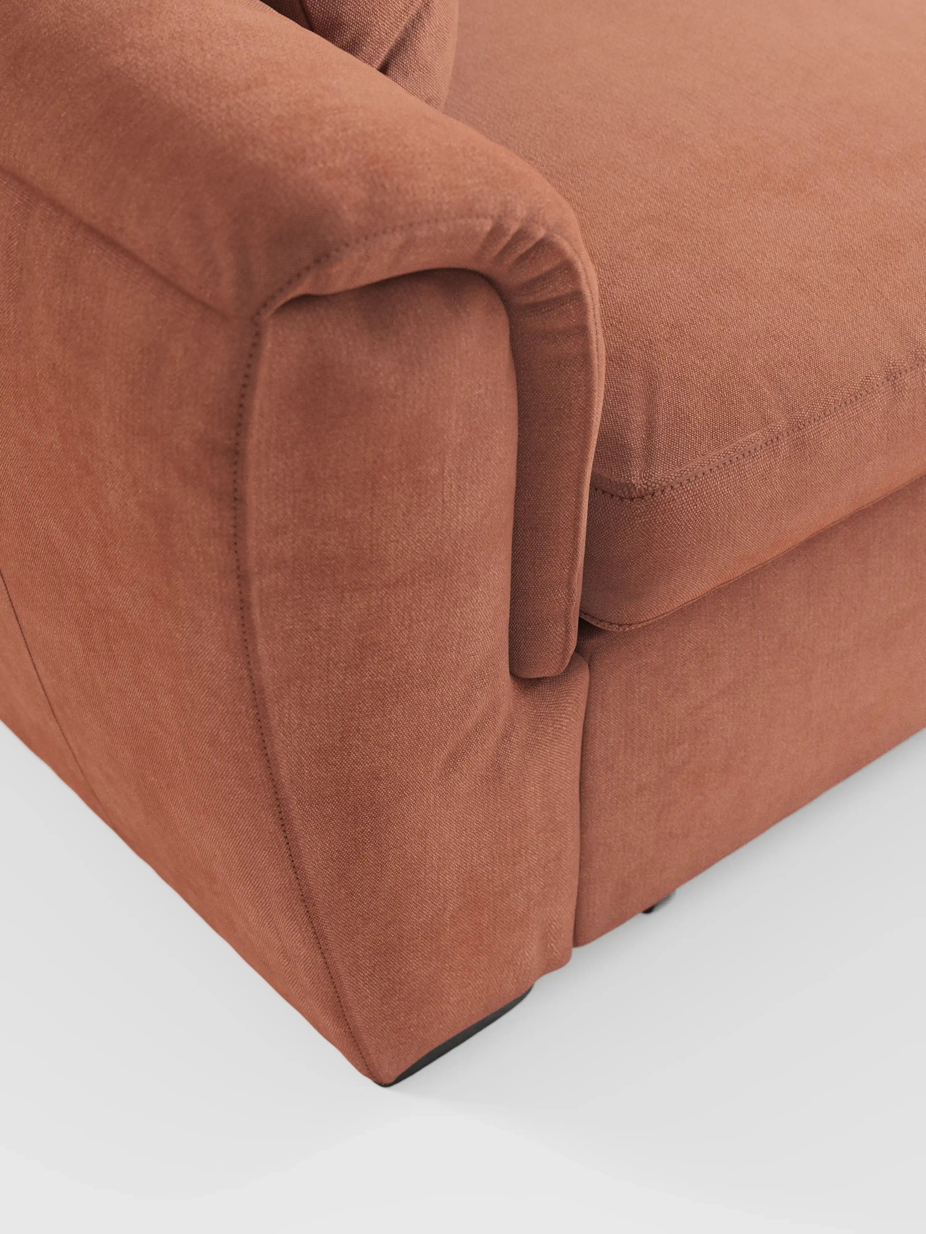 Primo piano dell'angolo del divano Titano a 2 posti estraibile con poggiatesta reclinabili in tessuto smacchiabile Oeko-Tex® di Divani.Store, che mette in risalto il bracciolo imbottito e il cuscino di seduta. Il rivestimento color ruggine presenta un aspetto morbido e materico, mentre le raffinate cuciture e le linee eleganti risaltano sullo sfondo bianco. Questo divano compatto e moderno è ideale per gli spazi abitativi più piccoli, offrendo al contempo raffinatezza e comfort.