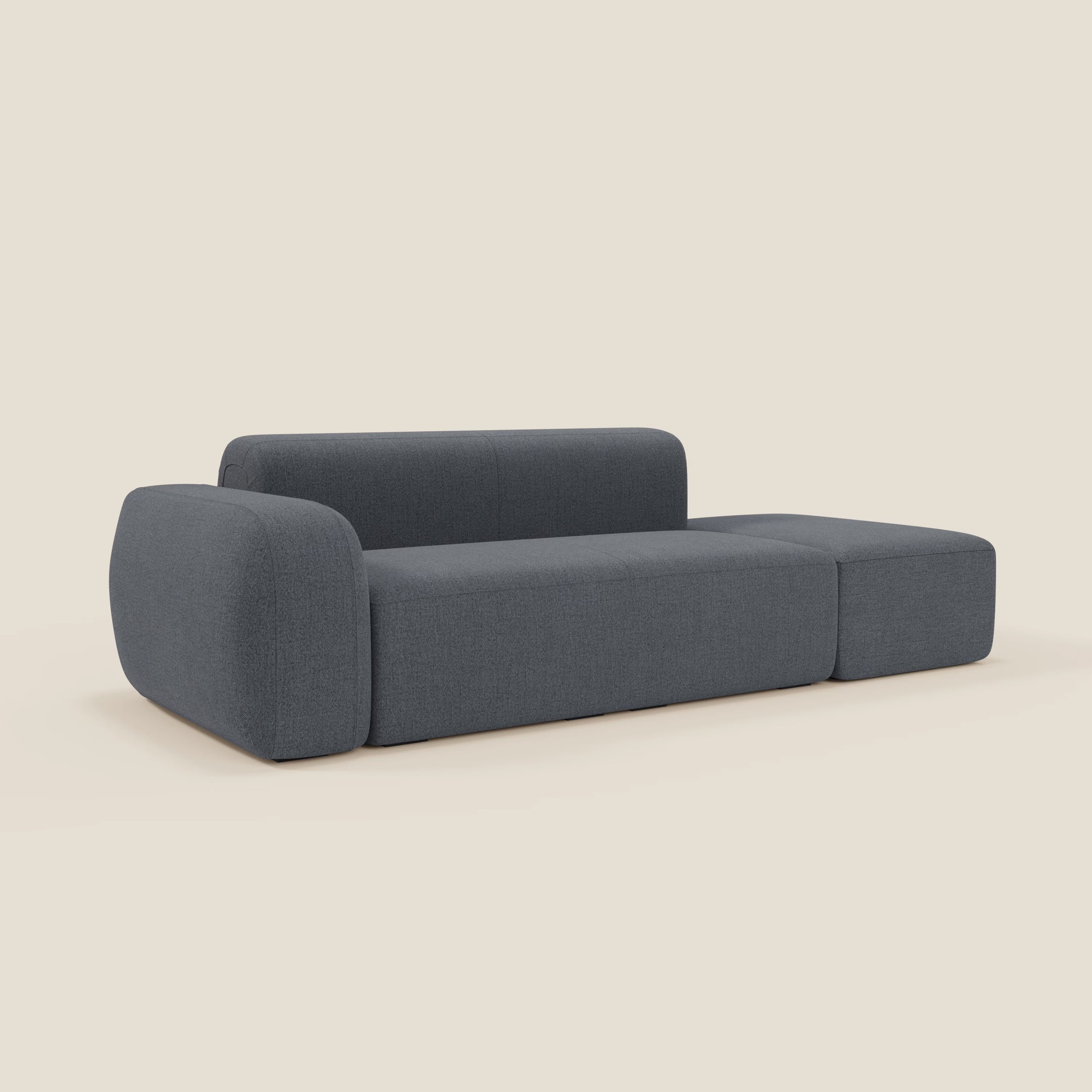 Il divano componibile BonBon di Divani.Store in tessuto T23 blu grafite smacchiabile è caratterizzato da un design minimalista con bordi arrotondati, schienale basso, ampi braccioli e pouf terminale, su uno sfondo bianco sporco.