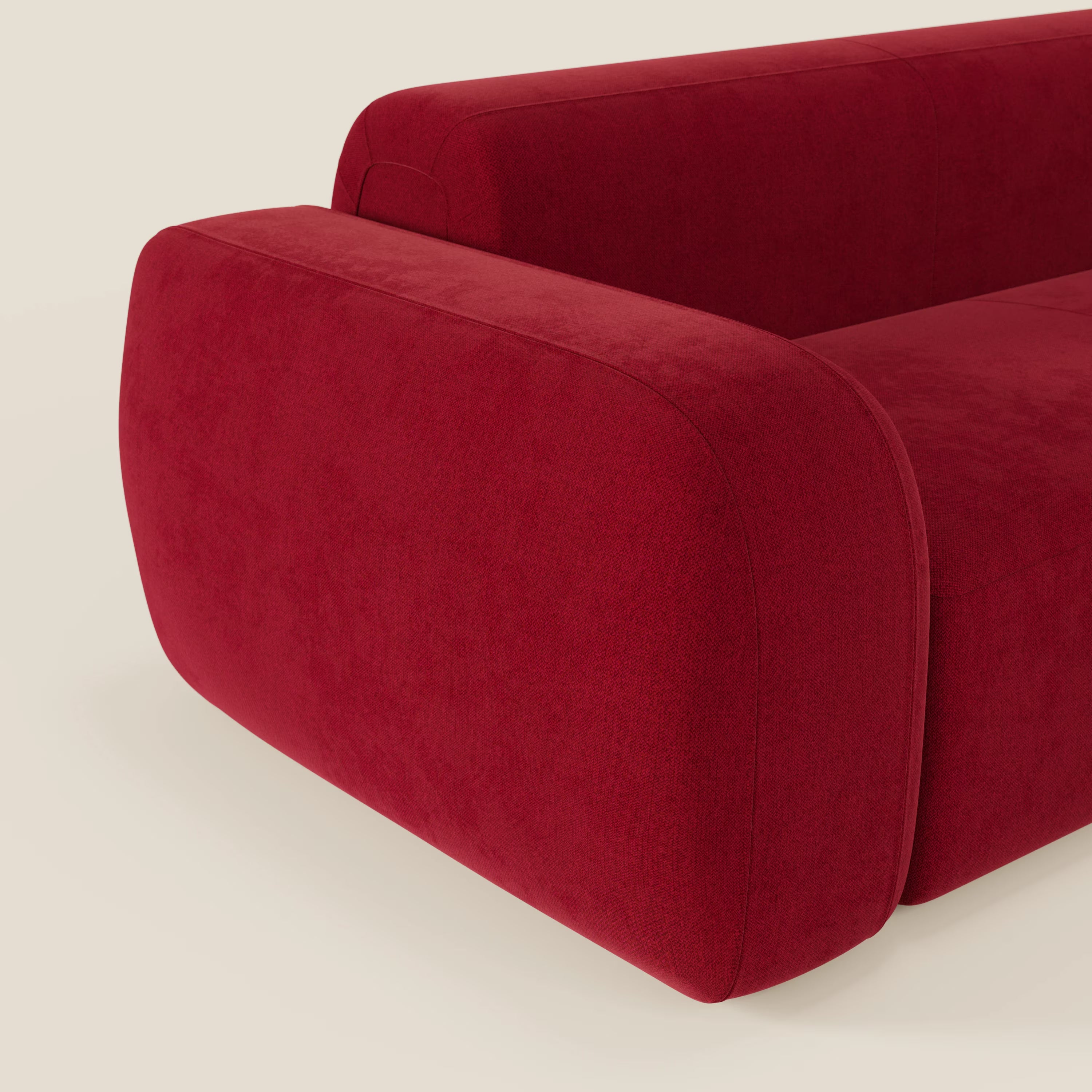 Primo piano del divano componibile BonBon Divano di Divani.Store in tessuto smacchiabile T23 bordeaux, con bordi arrotondati, braccioli e seduta spessi e pouf terminale opzionale su sfondo bianco sporco.
