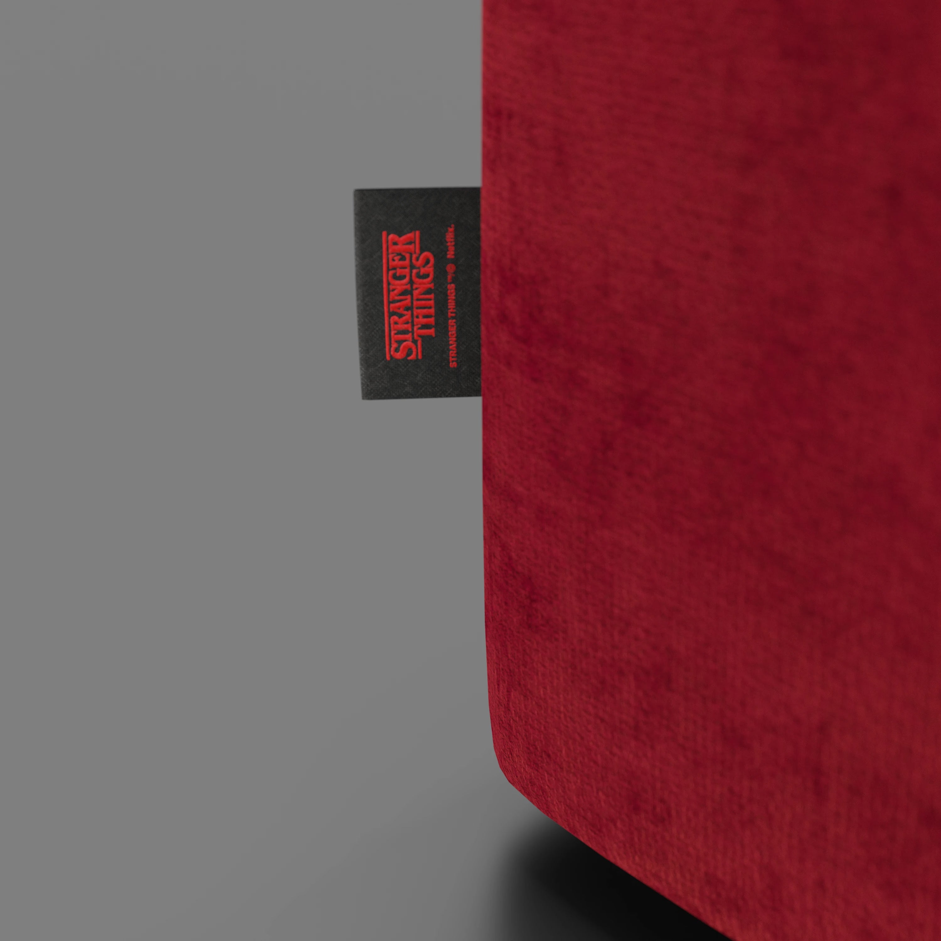 Primo piano del pouf Cube Stranger Things Edition di Divani.Store, caratterizzato da un tessuto rosso antimacchia e da una piccola etichetta nera con il logo rosso di "Stranger Things", su uno sfondo grigio.