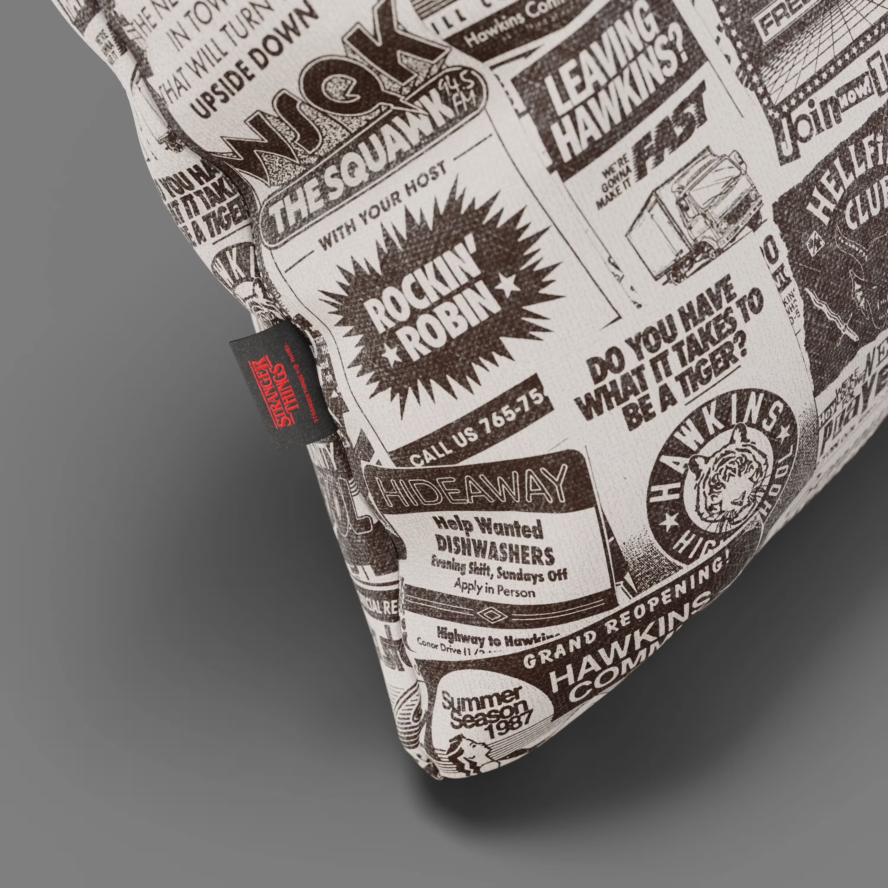 Un primo piano del Cuscino Divani.Store Hawkins Post Stranger Things - Limited Edition 45x45 cm, caratterizzato da stampe di giornali retrò in bianco e nero con annunci e titoli come "ROCKIN' ROBIN" e "HELP WANTED DISHWASHERS".