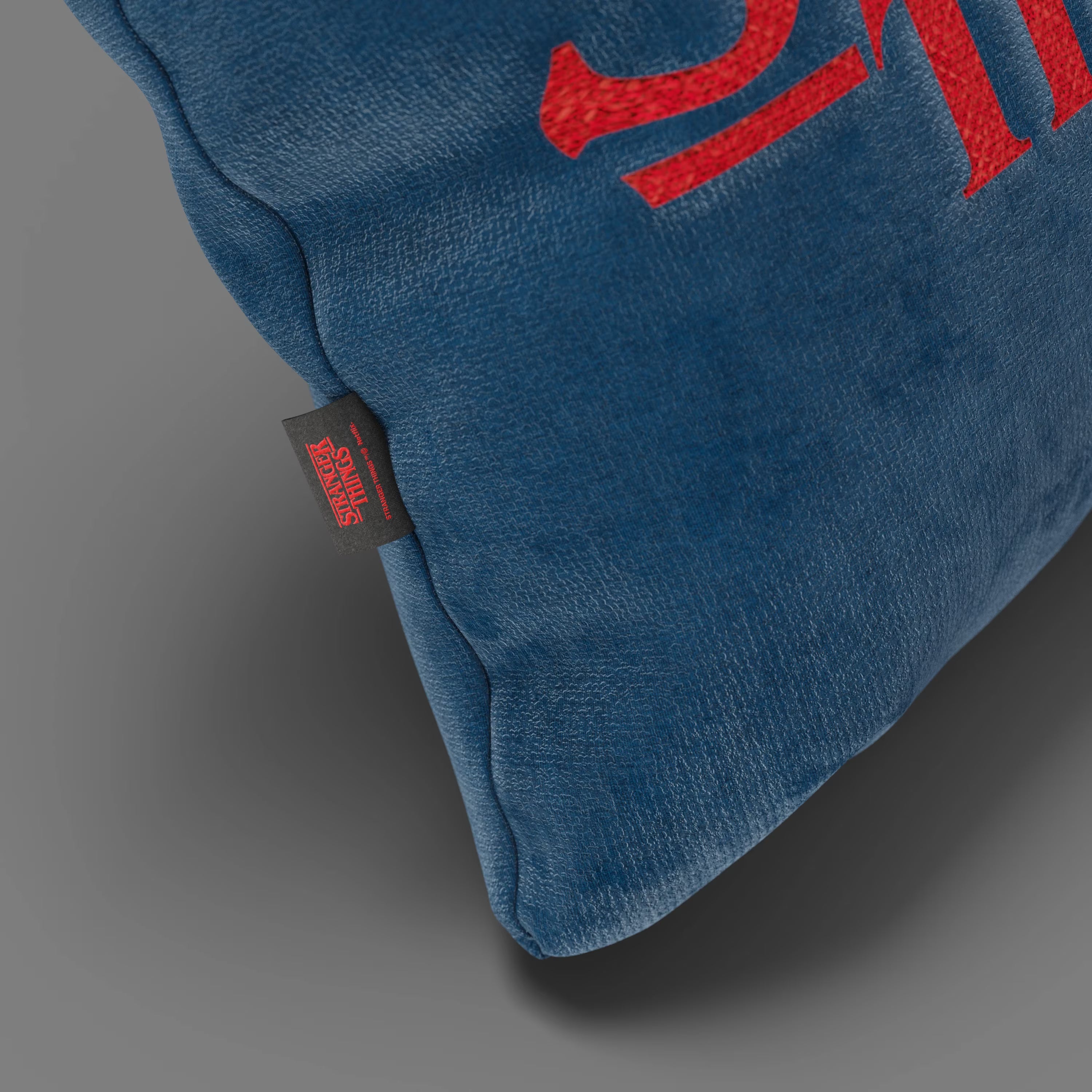 Primo piano del Cuscino Divani.Store Stranger Things Limited Edition con Ricamo in tessuto blu smacchiabile (45x45), con testo rosso e targhetta nera "Stranger Things", su sfondo grigio.