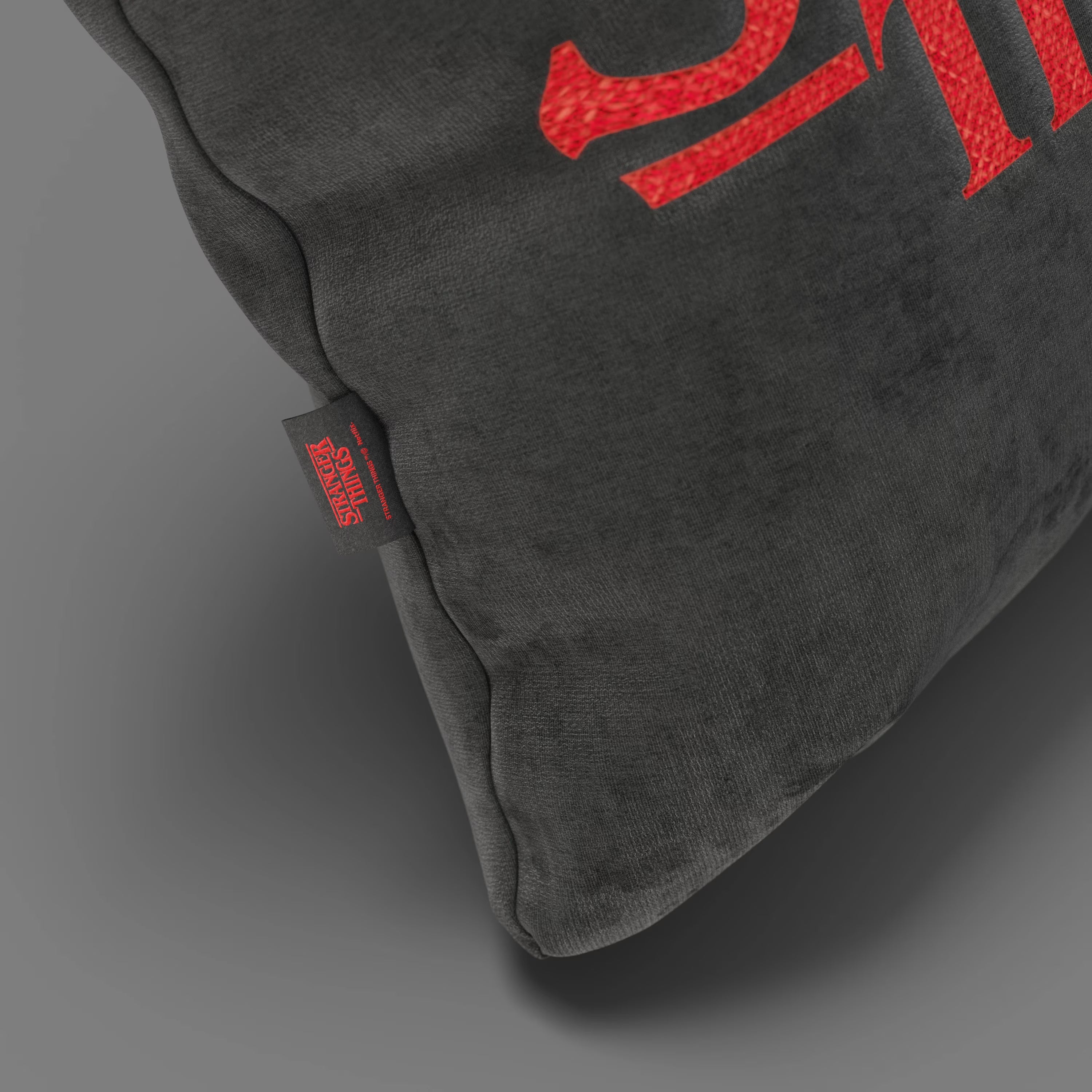 Primo piano del Cuscino Stranger Things Limited Edition con Ricamo in tessuto smacchiabile antracite (45x45) di Divani.Store, che mostra la sua texture sfocata, il dettaglio del testo rosso e l'etichetta nera di Netflix "Stranger Things" su uno sfondo grigio.