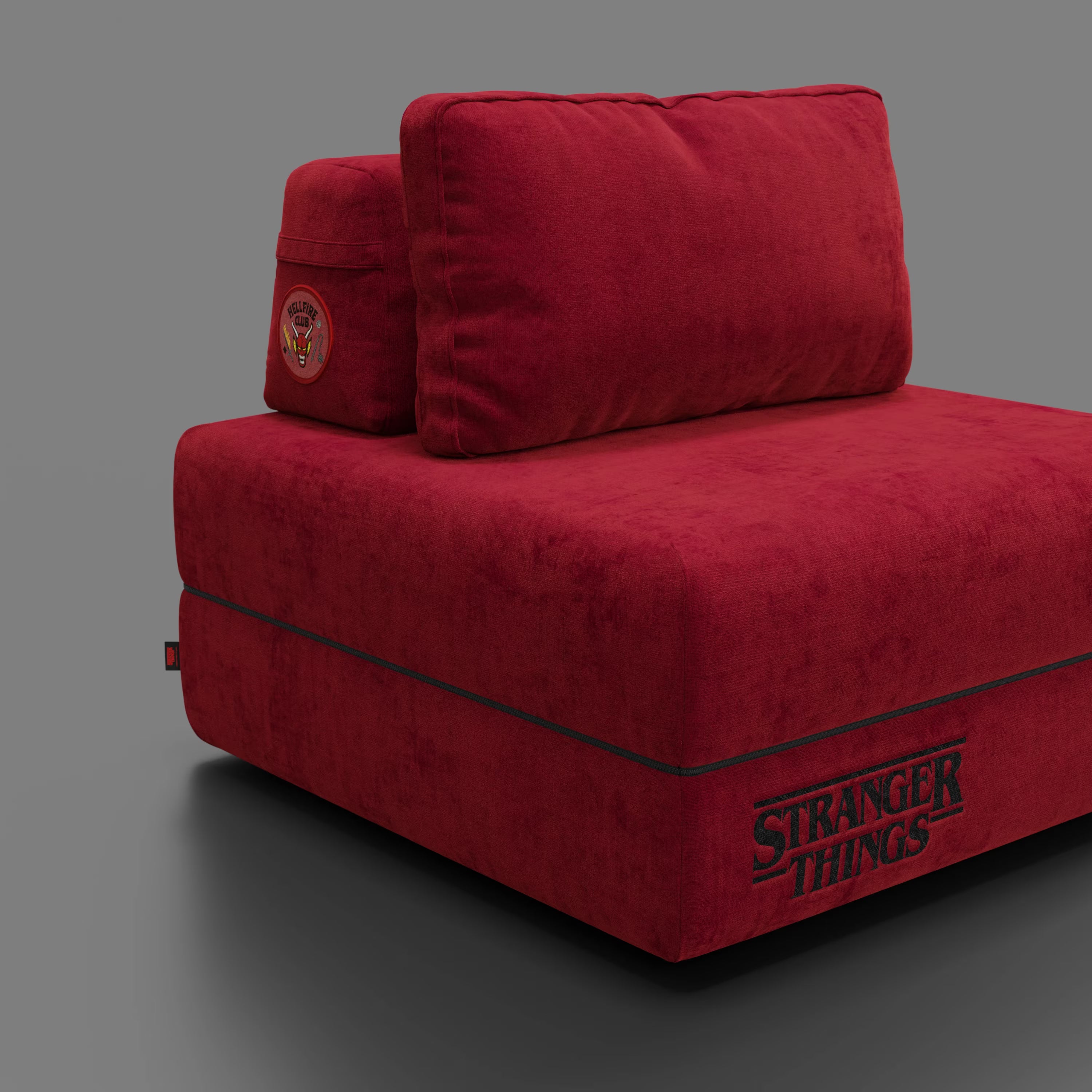 Una poltrona letto Rift rossa STRANGER THINGS Edition di Divani.Store, con materasso in memory foam da 22 cm e tessuto antimacchia, presenta una toppa "Hawkins High School" e il ricamo "Stranger Things Edition" su sfondo grigio.