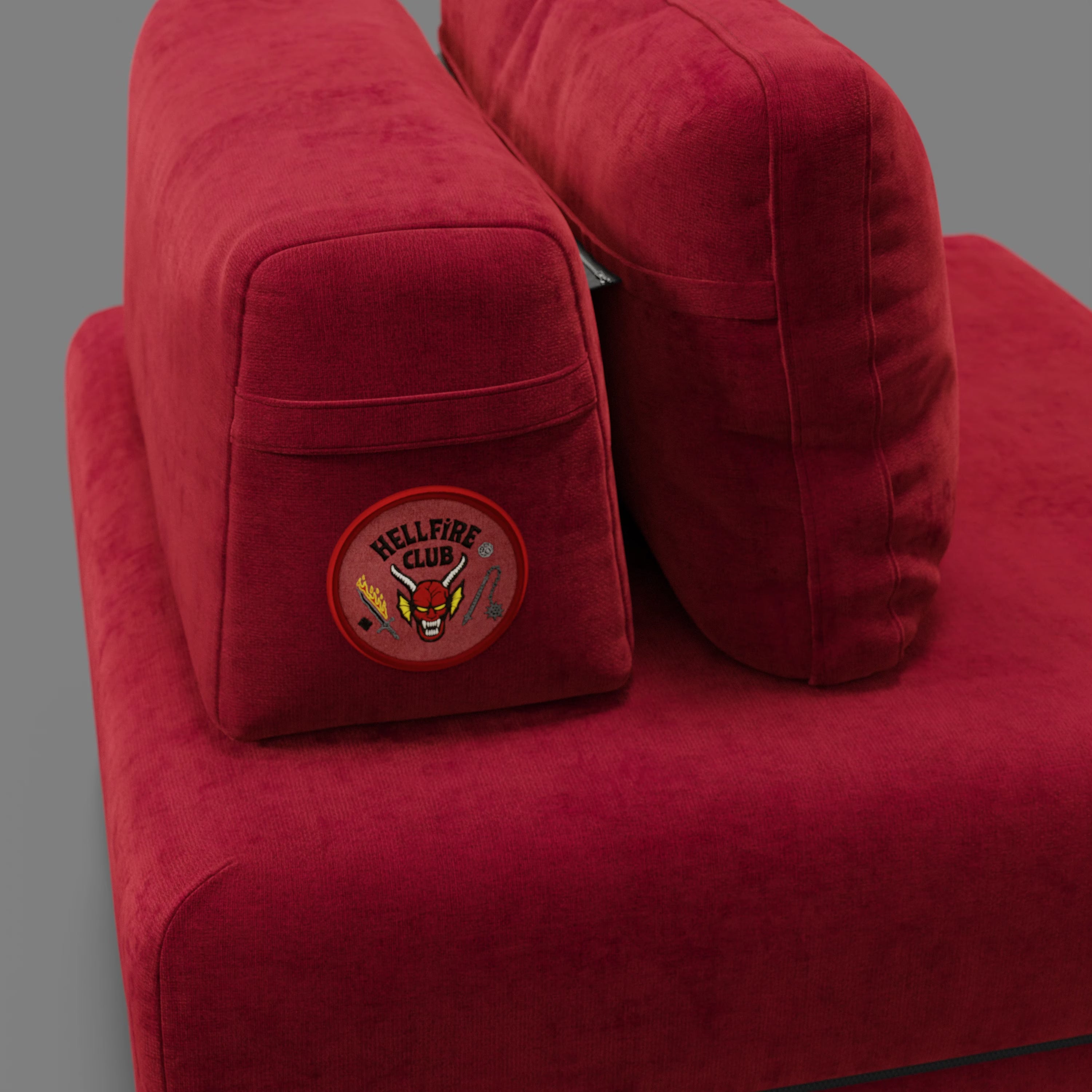 Primo piano della poltrona letto Rift STRANGER THINGS Edition di Divani.Store in rosso, con due cuscini verticali, di cui uno con patch circolare "Hellfire Club". Il divano ha un materasso in memory foam di 22 cm e un tessuto antimacchia, su uno sfondo grigio neutro.