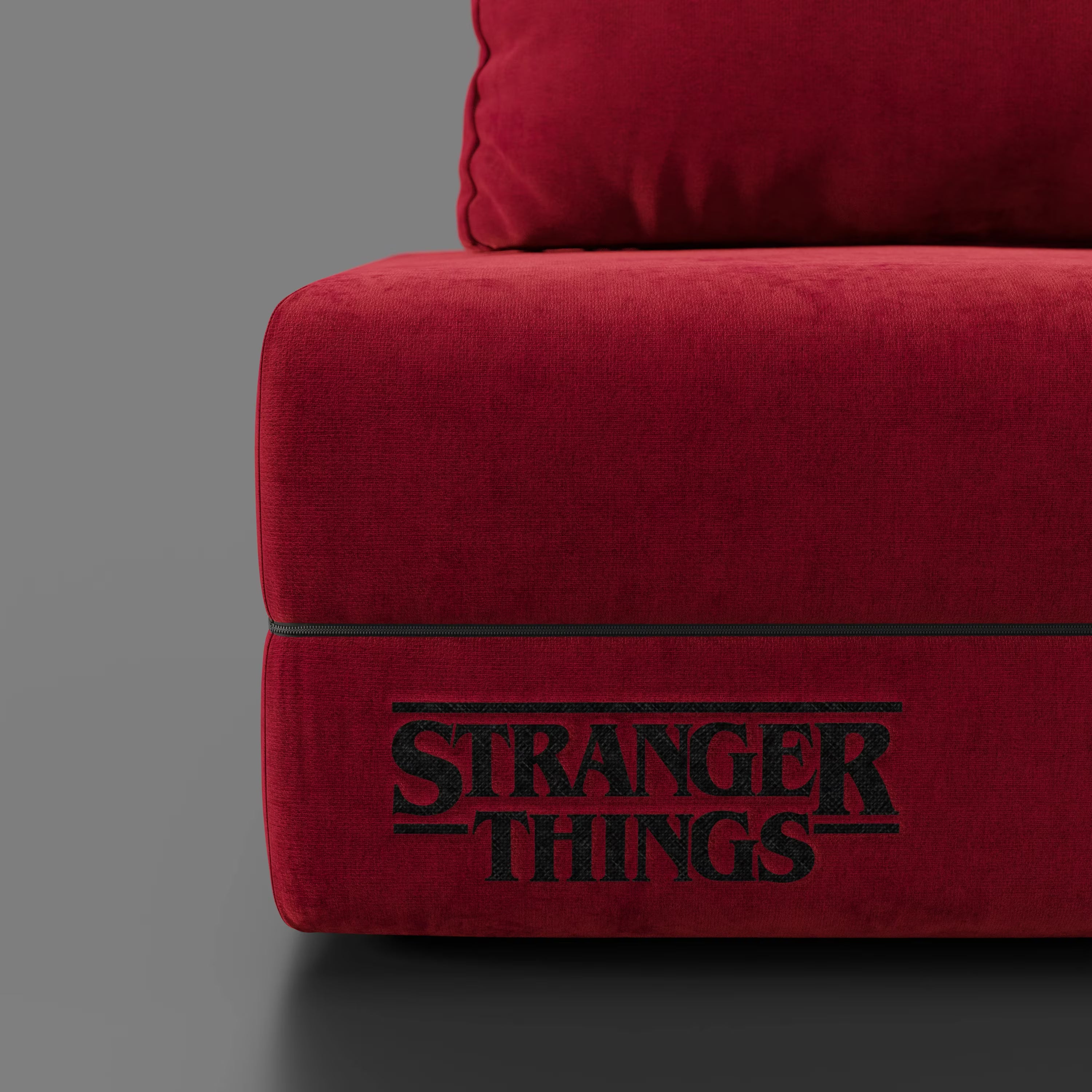 Primo piano della poltrona letto Rift STRANGER THINGS Edition di Divani.Store in tessuto rosso antimacchia con materasso in memory foam da 22 cm, che evidenzia l'esclusivo logo ricamato su uno sfondo grigio liscio.