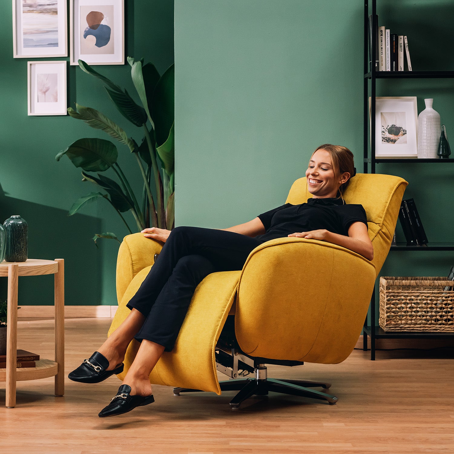 Una donna in nero si rilassa, sorridendo a occhi chiusi, sulla poltroncina EVA RELAX in morbido tessuto impermeabile T02 giallo di Divani.Store. La stanza ha pareti verdi, pavimento in legno, piante, opere d'arte incorniciate, un tavolo e una libreria in metallo nero con libri.