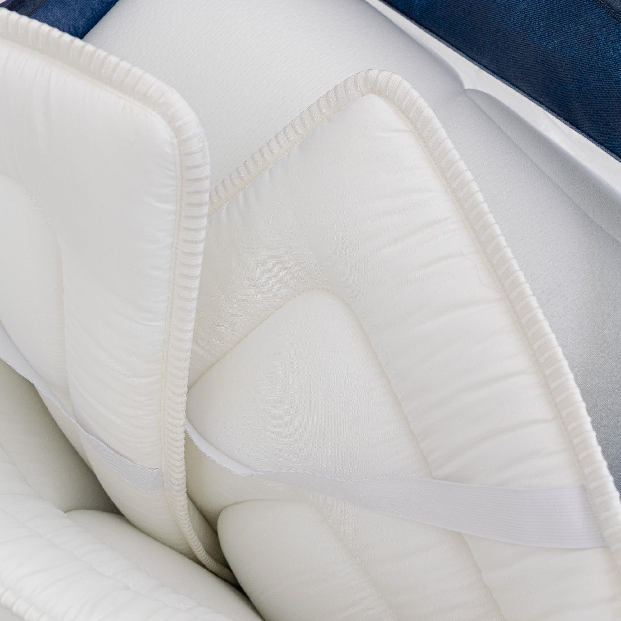 Primo piano di due coprimaterassi bianchi trapuntati, tra cui il Divani.Store KYROS-CLOUD Topper Luxury in Memory Foam H5 cm con Tecnologia CoolMax con bordi cuciti e cinturino elastico. Offrono un comfort ammortizzato; sullo sfondo appare un tessuto blu scuro.