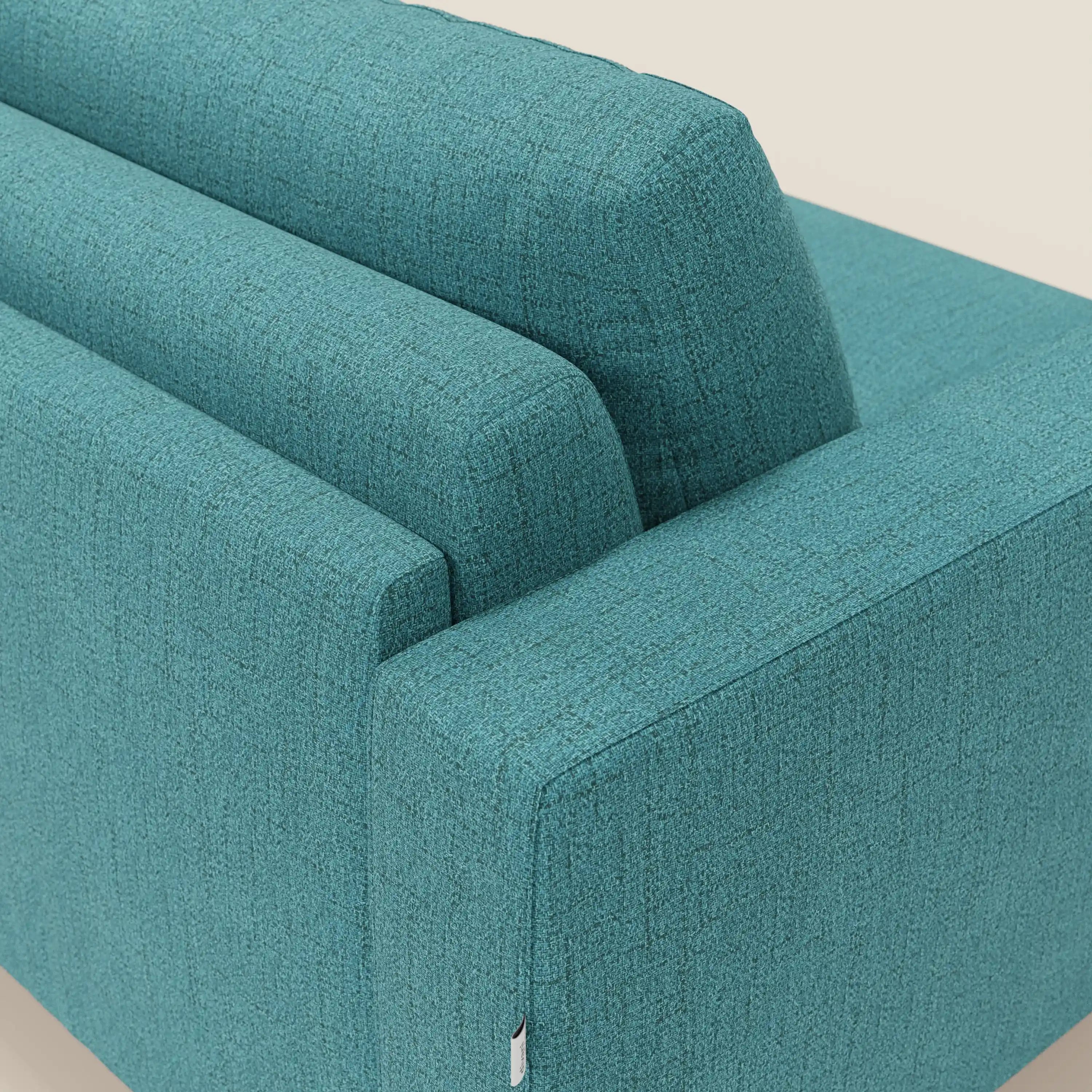 Una vista angolata e ravvicinata del letto matrimoniale Alaya Divano di Divani.Store in tessuto impermeabile T03 azzurro mette in evidenza i morbidi cuscini, lo schienale, il bracciolo e la trama intrecciata. Una targhetta bianca è visibile sulla cucitura su uno sfondo bianco sporco.
