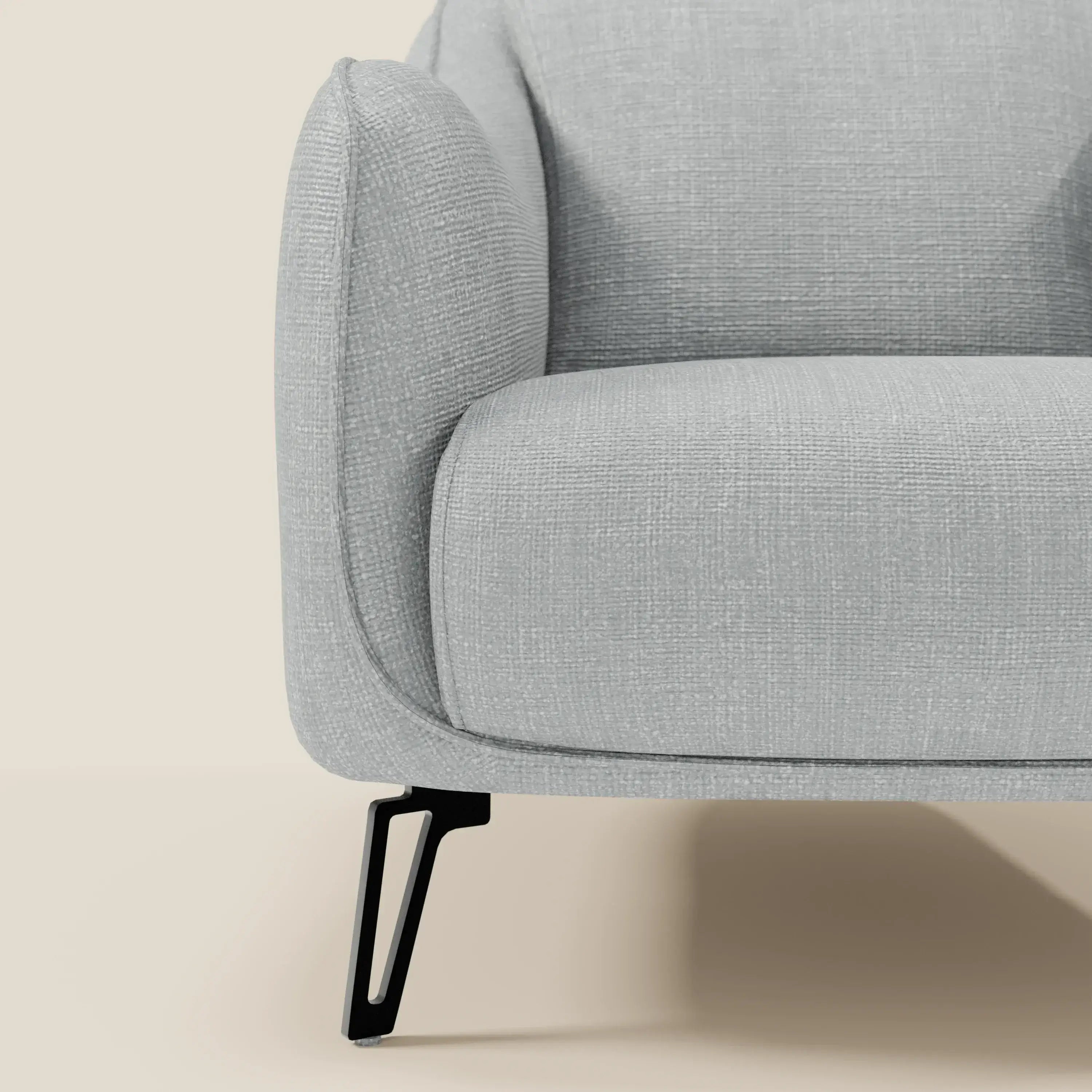 Primo piano della poltroncina Avela di Divani.Store in morbido tessuto impermeabile T06 ghiaccio, con bracciolo arrotondato, imbottitura testurizzata, elegante gamba in metallo nero e un design contemporaneo su sfondo beige.