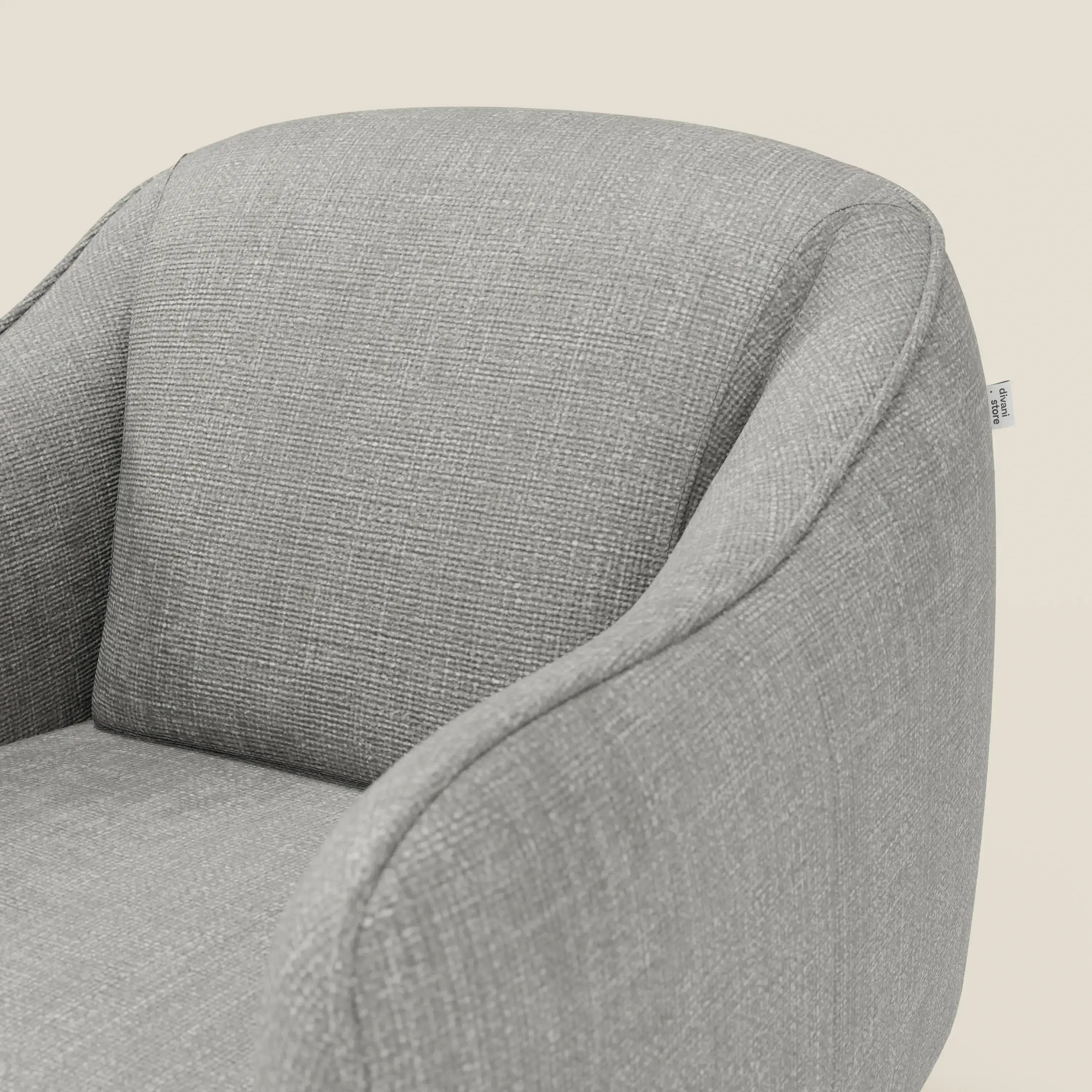 Primo piano di Avela Poltroncina di Divani.Store, una poltrona dal comfort senza tempo in morbido tessuto impermeabile grigio T06. Lo schienale curvo e imbottito e i braccioli garantiscono il comfort; una piccola etichetta è visibile su uno sfondo bianco sporco.