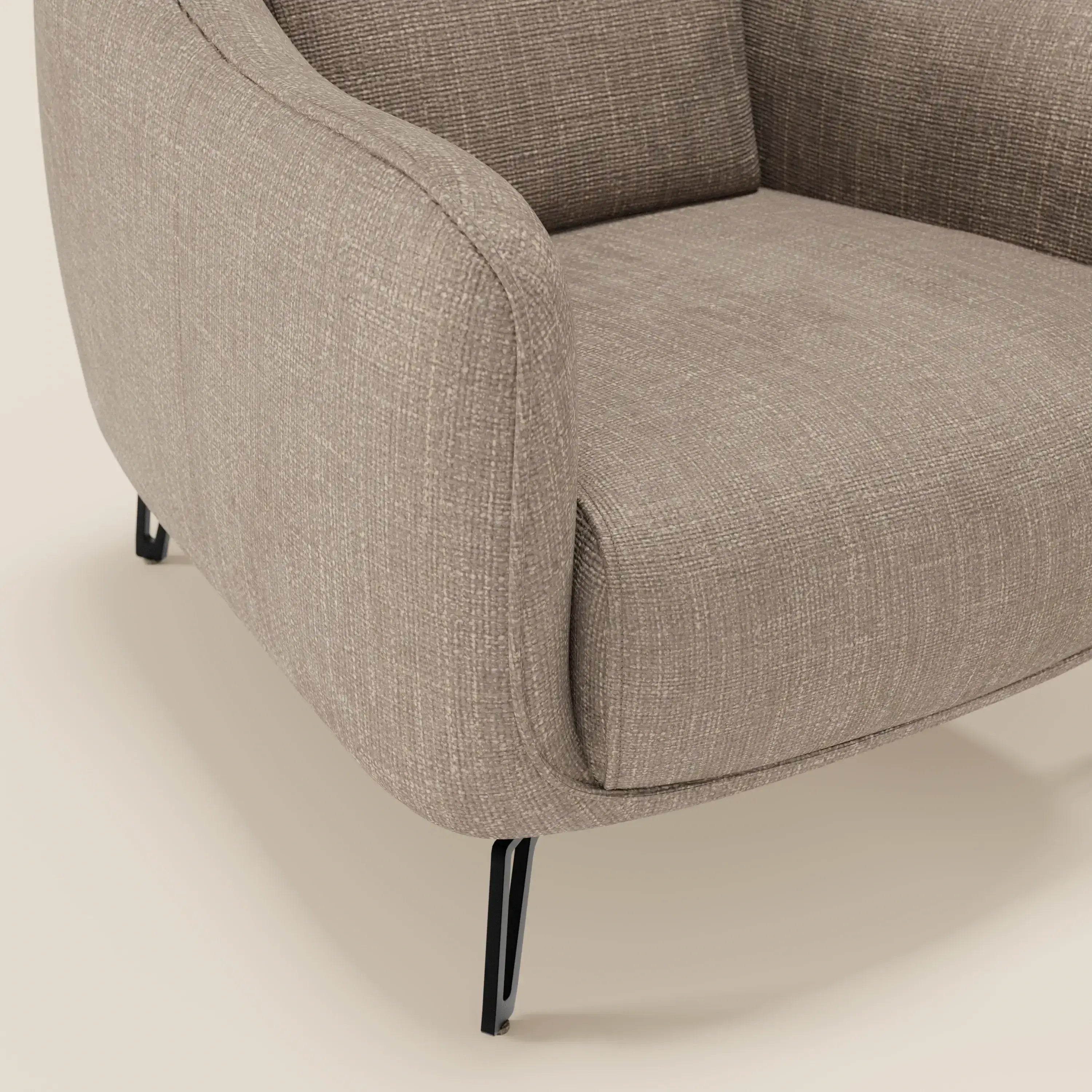 Primo piano di Avela Poltroncina di Divani.Store, caratterizzata da un morbido rivestimento in tessuto tortora T06 impermeabile, bordi arrotondati e sottili gambe in metallo nero. Il design è liscio e dolcemente curvo, su uno sfondo beige chiaro.