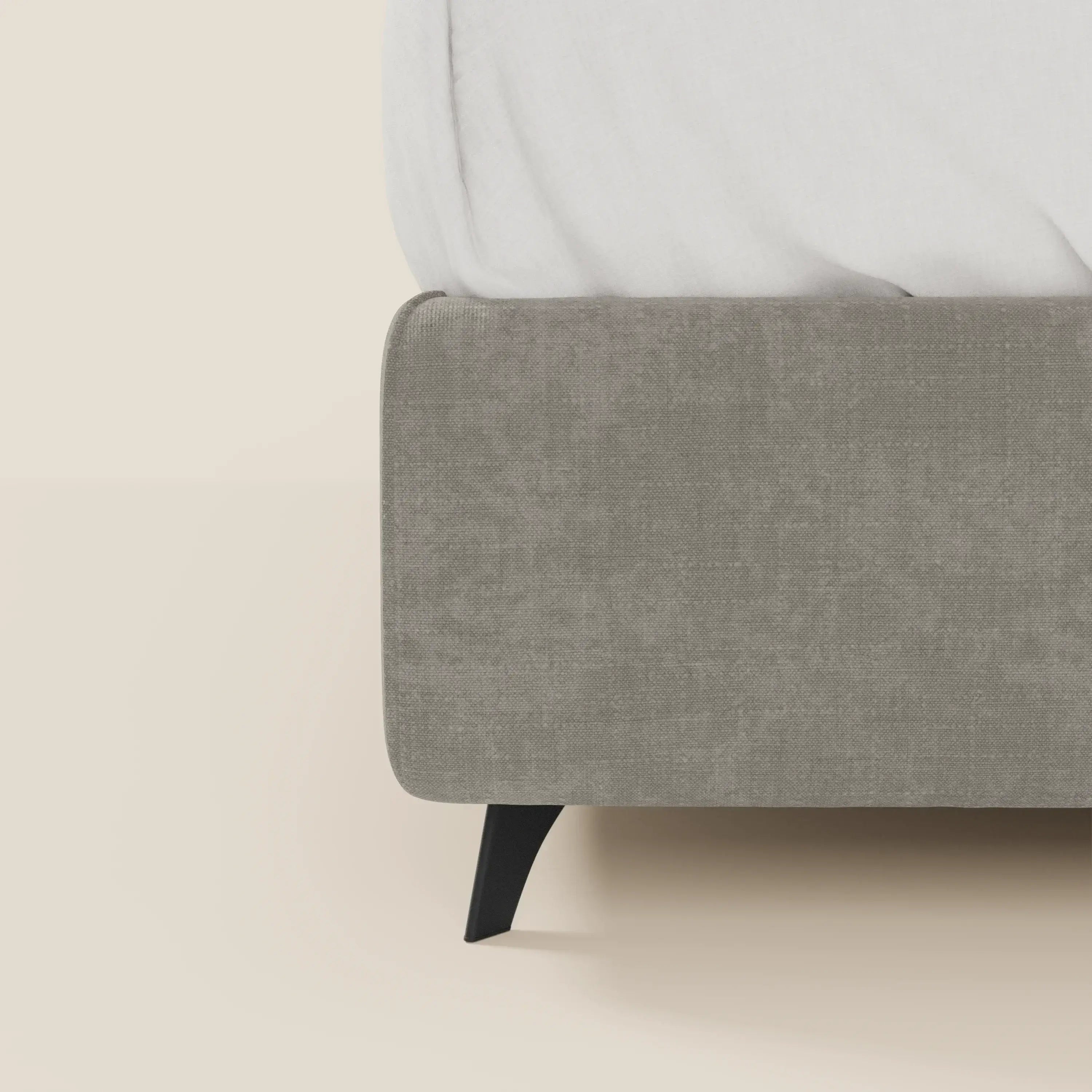 Primo piano del letto contenitore Bogart Edizione Grande Fratello di Letti.store, in tessuto T22 antimacchia grigio chiaro, sfoderabile e con gamba angolare nera. Il letto e il materasso sono visibili solo in parte, la biancheria è bianca su sfondo beige.