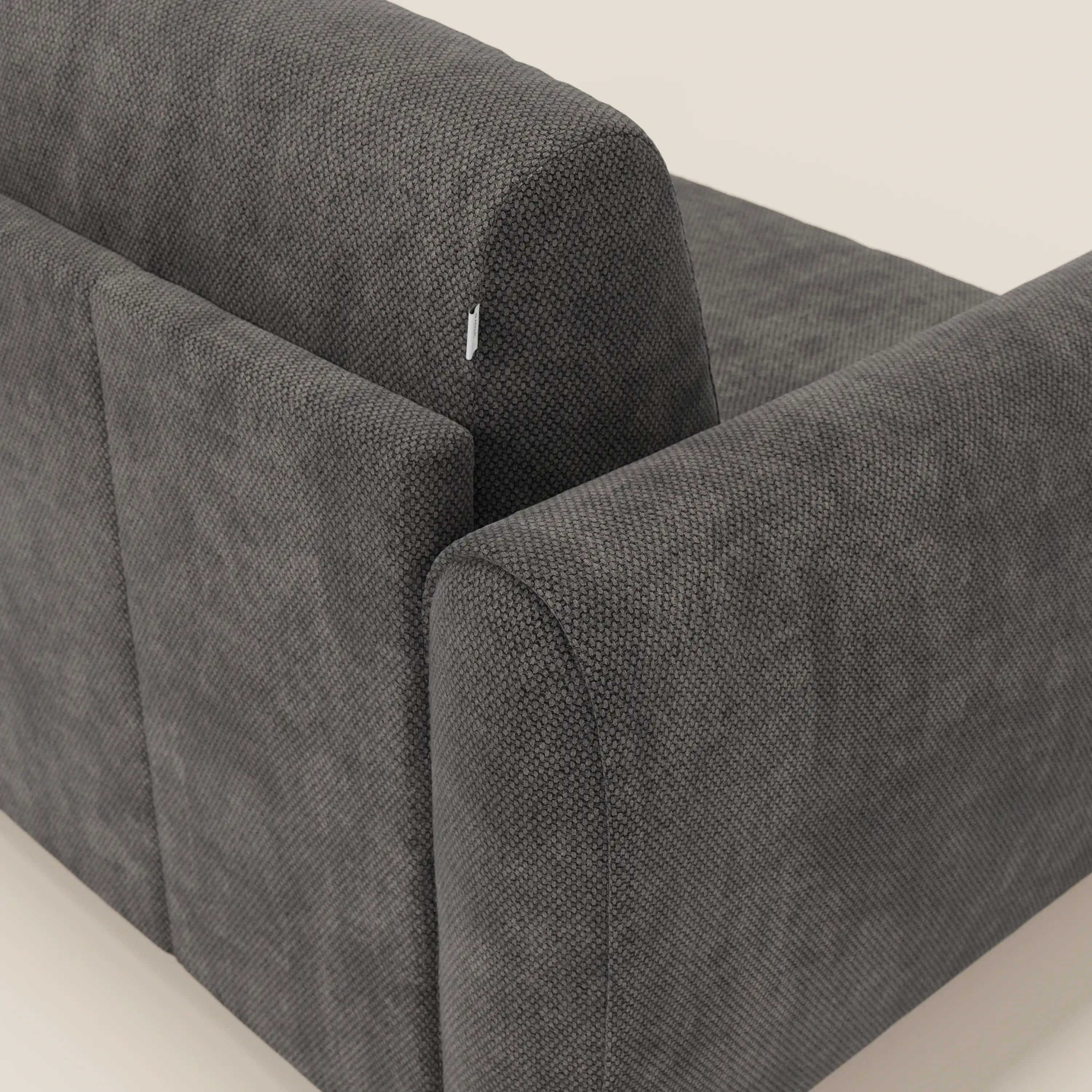 Un primo piano di Kaleo Divano letto moderno di Divani.Store in tessuto impermeabile T26 antracite mostra il rivestimento testurizzato, il bracciolo e lo schienale arrotondati, oltre a una piccola etichetta in tessuto bianco vicino al bordo superiore dello schienale su uno sfondo chiaro.