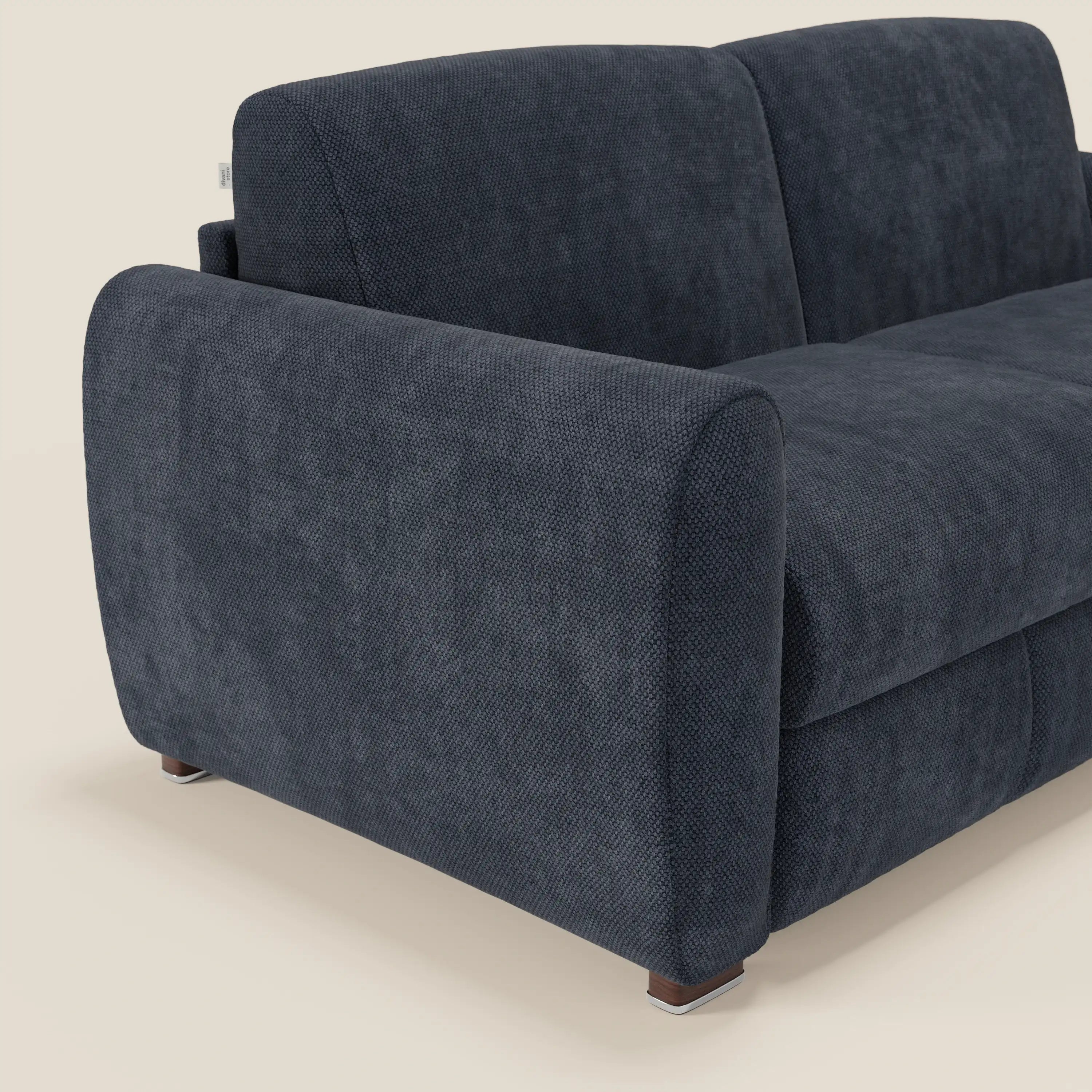 Un primo piano angolato del divano letto moderno Kaleo di Divani.Store in raffinato tessuto impermeabile T26 blu navy, caratterizzato da una morbida imbottitura, un ampio bracciolo arrotondato e corte gambe quadrate in legno con tappi in metallo su uno sfondo chiaro.