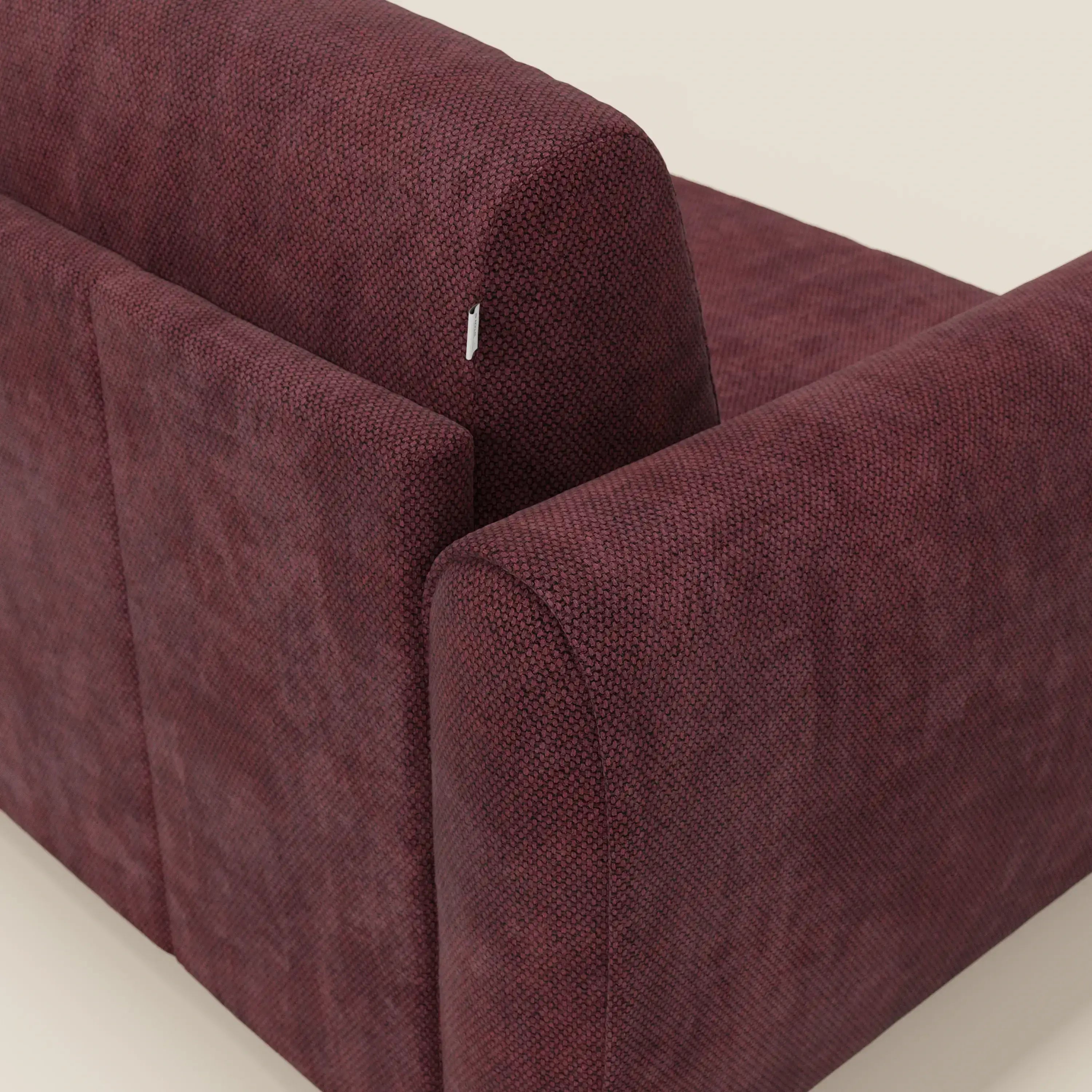 Primo piano del divano letto moderno Kaleo di Divani.Store, in raffinato tessuto impermeabile T26 bordeaux prugna, che mostra lo schienale e il bracciolo con una sottile texture e una piccola etichetta bianca, su uno sfondo bianco sporco.
