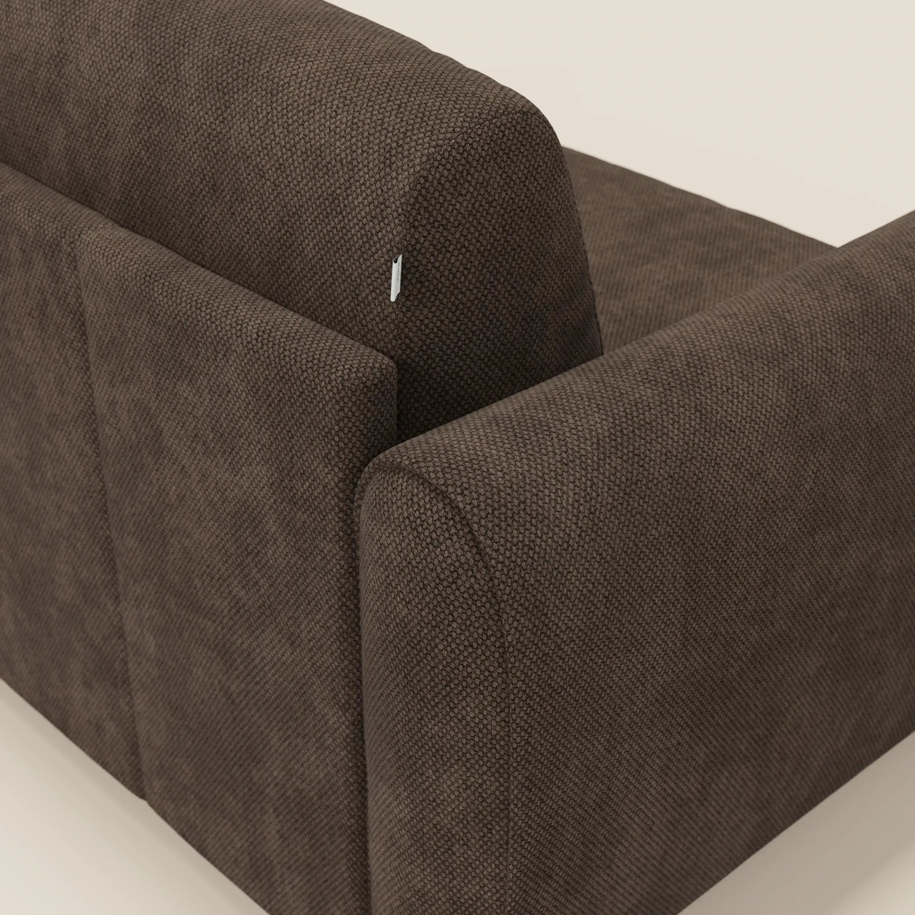 Un primo piano del divano letto moderno Kaleo di Divani.Store in tessuto impermeabile T26 marrone ne evidenzia le linee pulite e il design contemporaneo, con una piccola targhetta bianca sul cuscino e uno sfondo beige semplice.