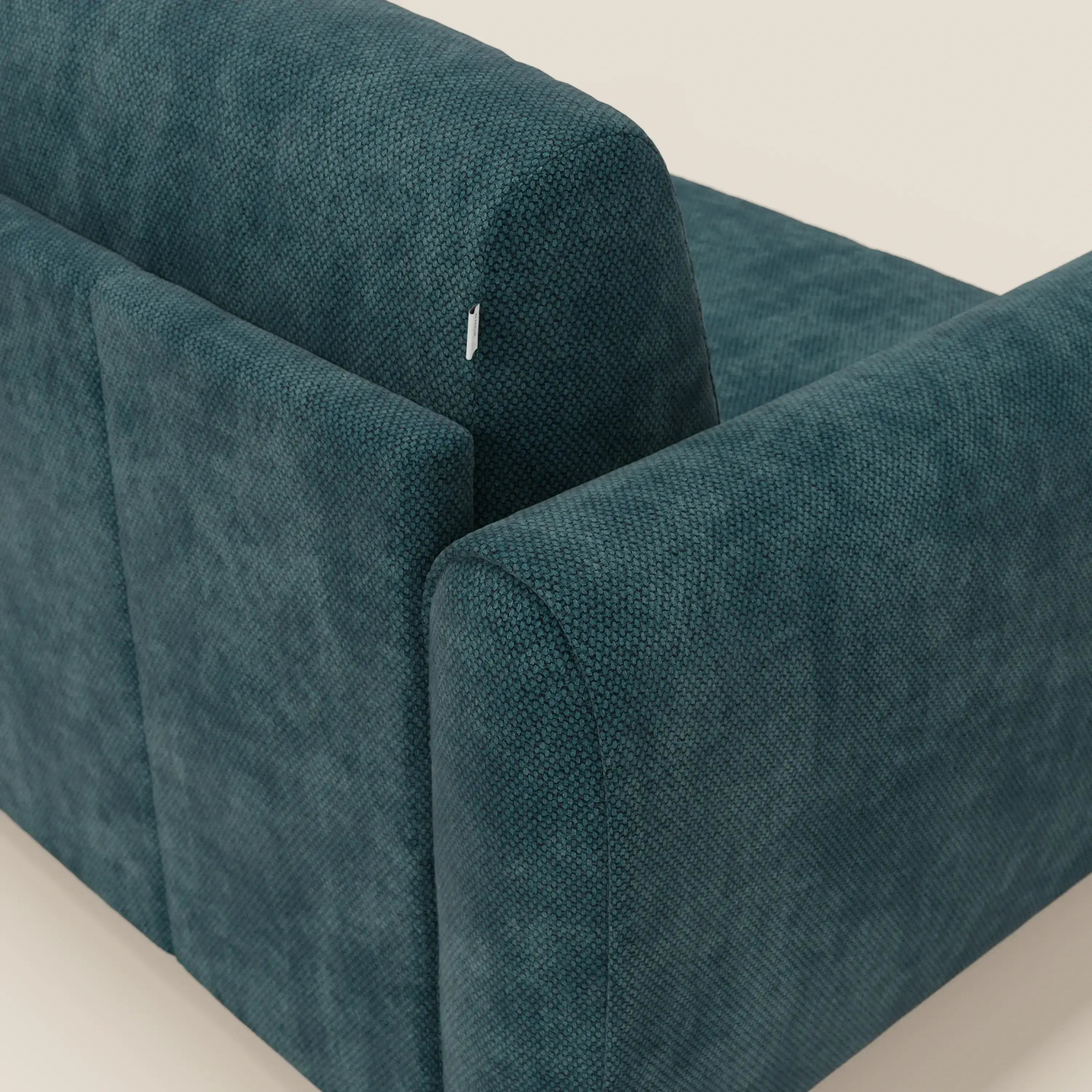 Un primo piano di Kaleo Divano letto moderno di Divani.Store in tessuto impermeabile T26 petrolio mostra il rivestimento testurizzato e una piccola etichetta in tessuto bianco sullo schienale superiore, su uno sfondo beige chiaro per un look contemporaneo.