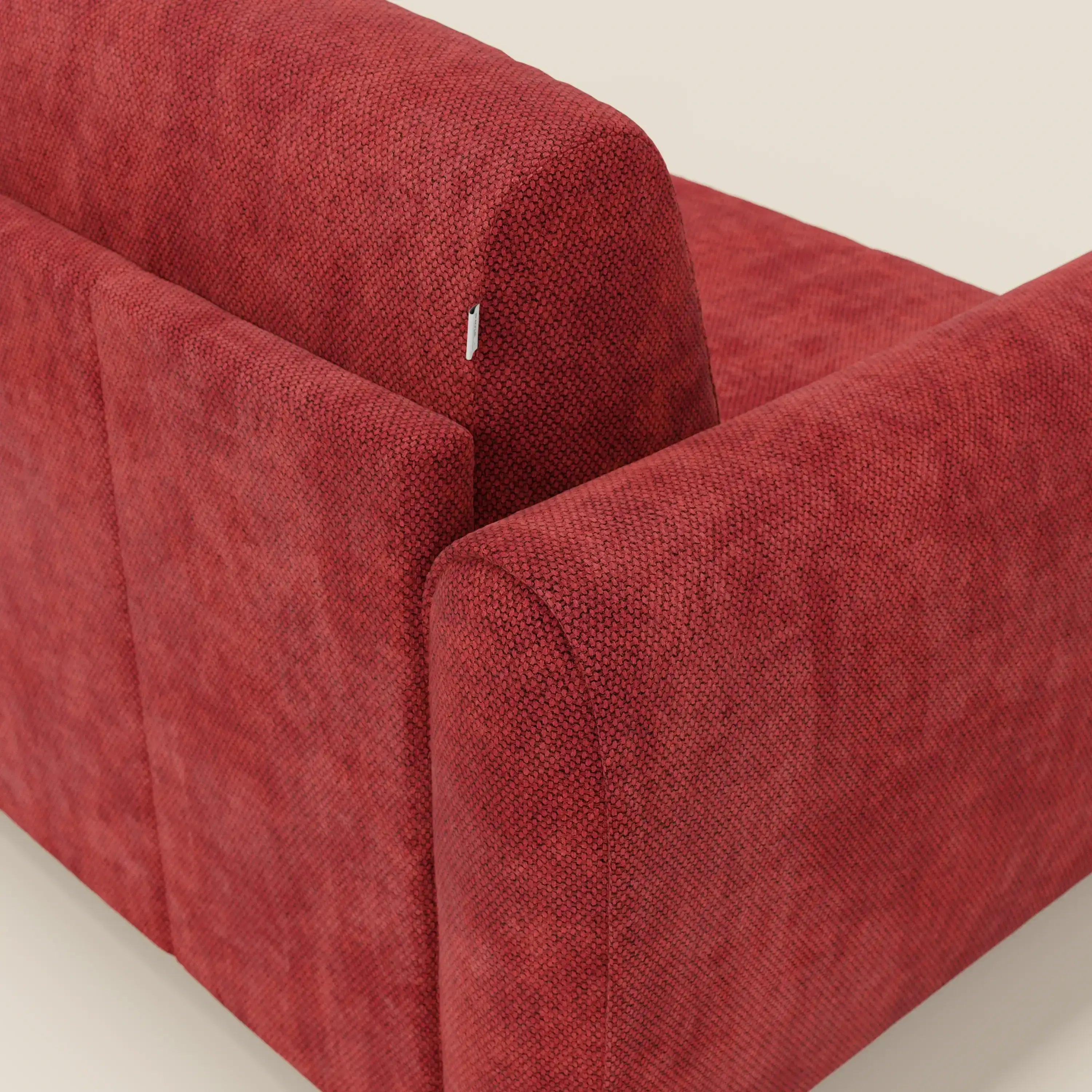 Un primo piano del Kaleo Divano letto moderno di Divani.Store, in tessuto impermeabile rosso T26, mette in evidenza la sua consistenza felpata e dettagliata e una piccola etichetta bianca sullo schienale, su uno sfondo beige chiaro.