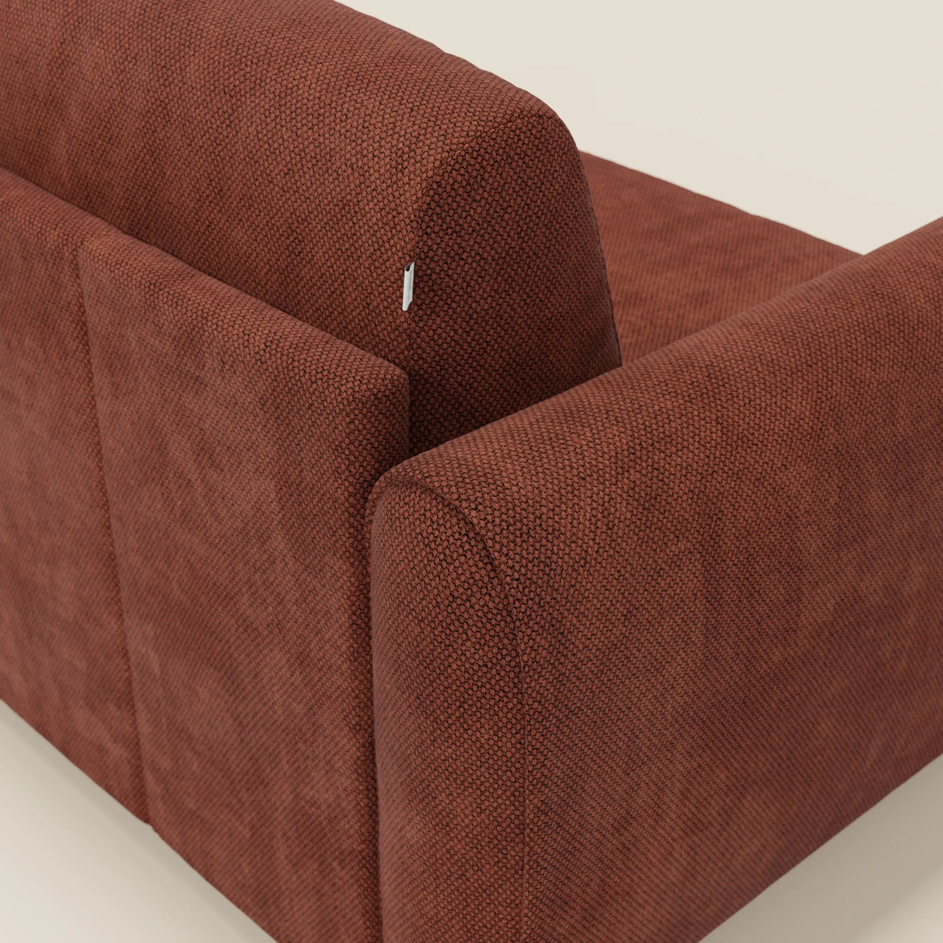 Primo piano dello schienale e del bracciolo di Kaleo Divano letto moderno di Divani.Store in tessuto arancione ruggine T26 testurizzato e impermeabile. Una piccola etichetta di tessuto è visibile vicino al bordo superiore, su uno sfondo neutro e chiaro.