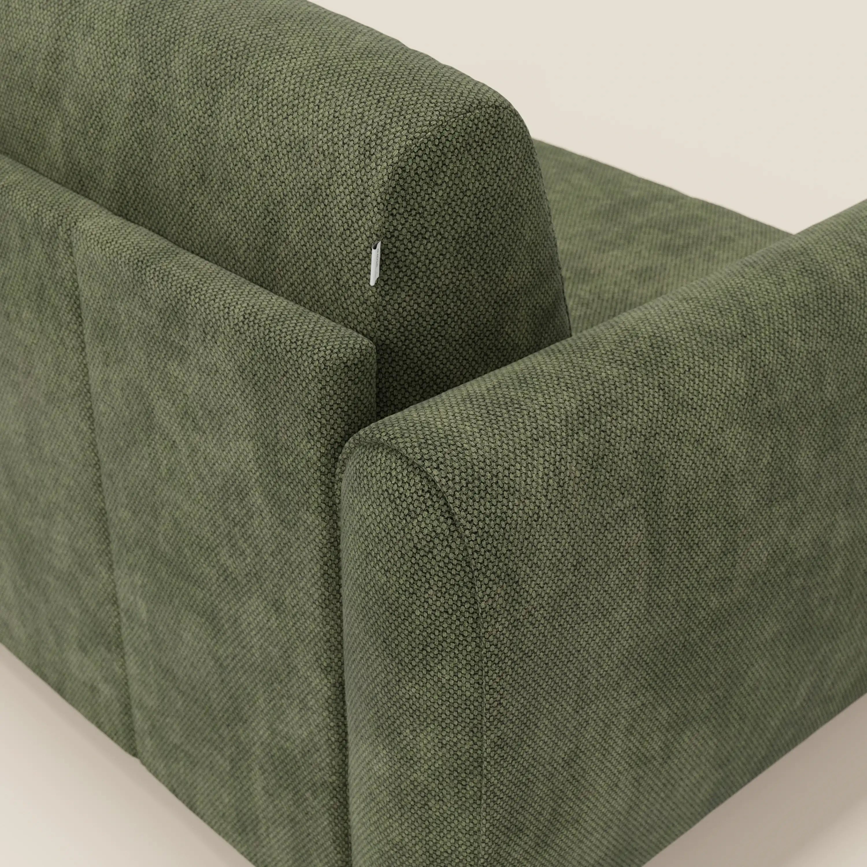 Primo piano dello schienale e del bracciolo del divano letto moderno Divani.Store Kaleo in raffinato tessuto impermeabile T26 verde muschio, che mostra il suo design contemporaneo, le linee pulite, il rivestimento testurizzato e una piccola etichetta bianca sullo schienale superiore. Sfondo beige chiaro.