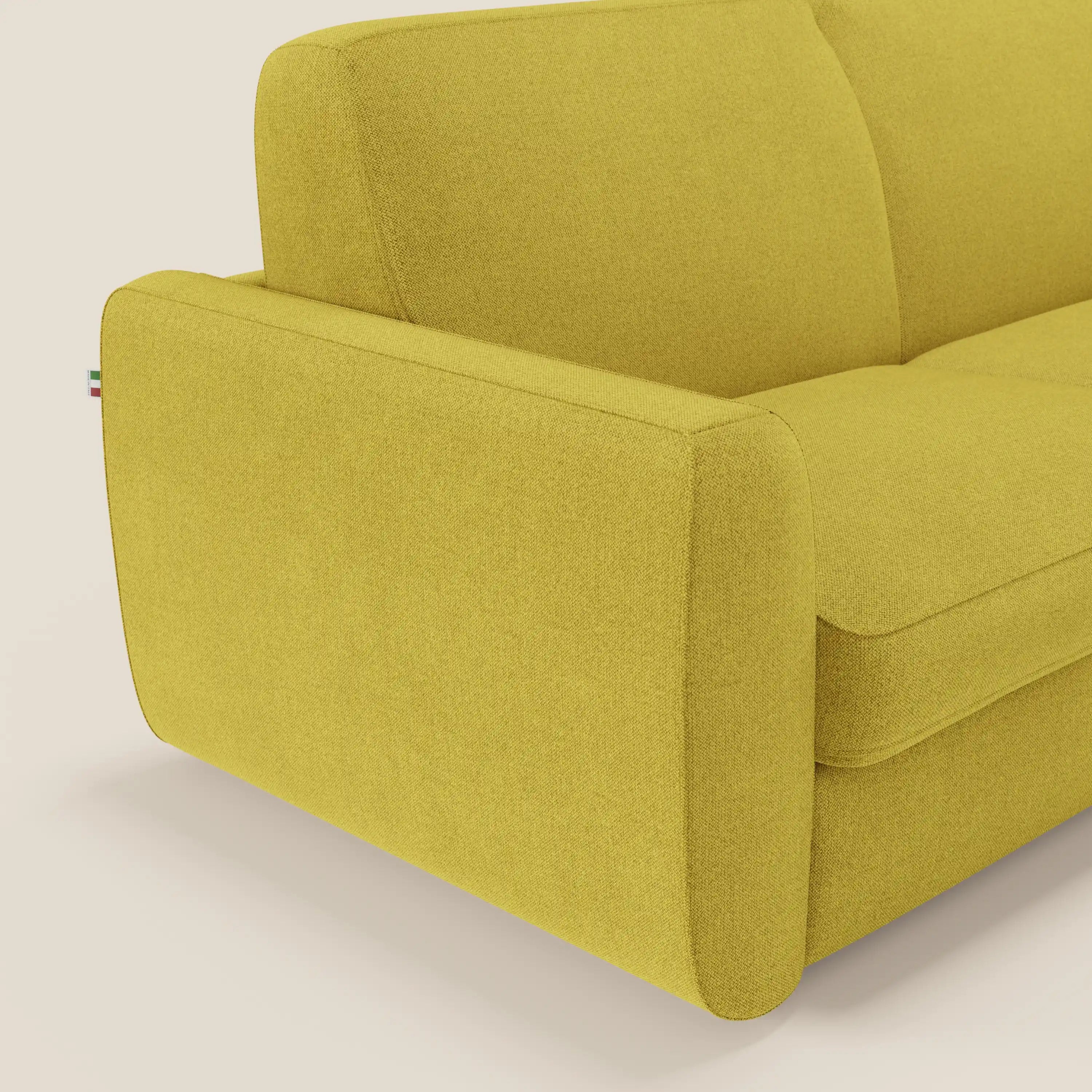 Un primo piano del lato sinistro del divano letto sfoderabile Leonida di Divani.Store in tessuto Smacchiabile T23 giallo sole mostra il bracciolo, la seduta, lo schienale e la targhetta con i colori della bandiera italiana, accennando al materasso H18 cm e al vano portaguanciali.