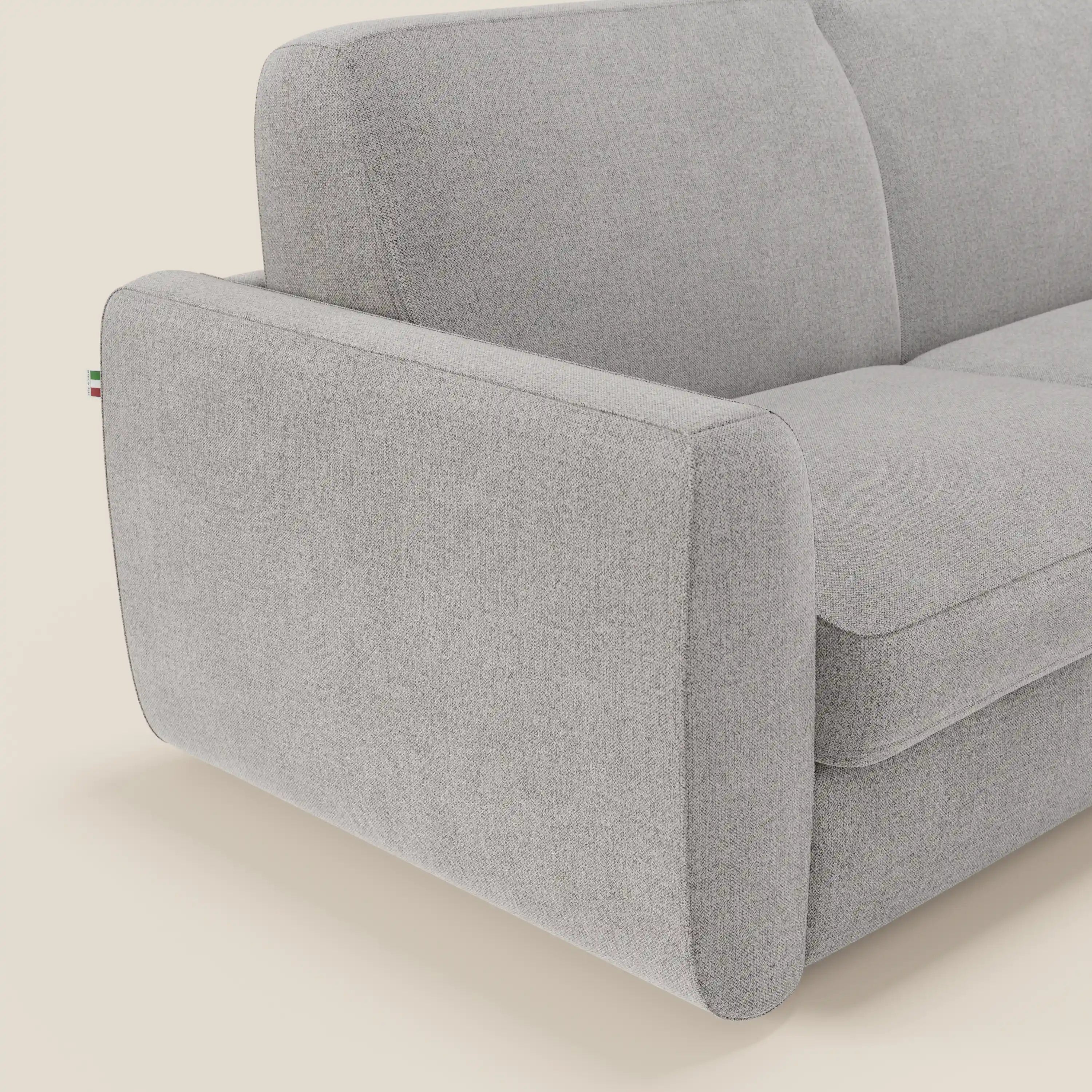 Un primo piano del divano letto sfoderabile Leonida di Divani.Store, in tessuto smacchiabile T23 grigio, evidenzia il bracciolo sinistro con la targhetta della bandiera italiana. Il fondo bianco sporco accentua il design moderno e il materasso H18 cm.