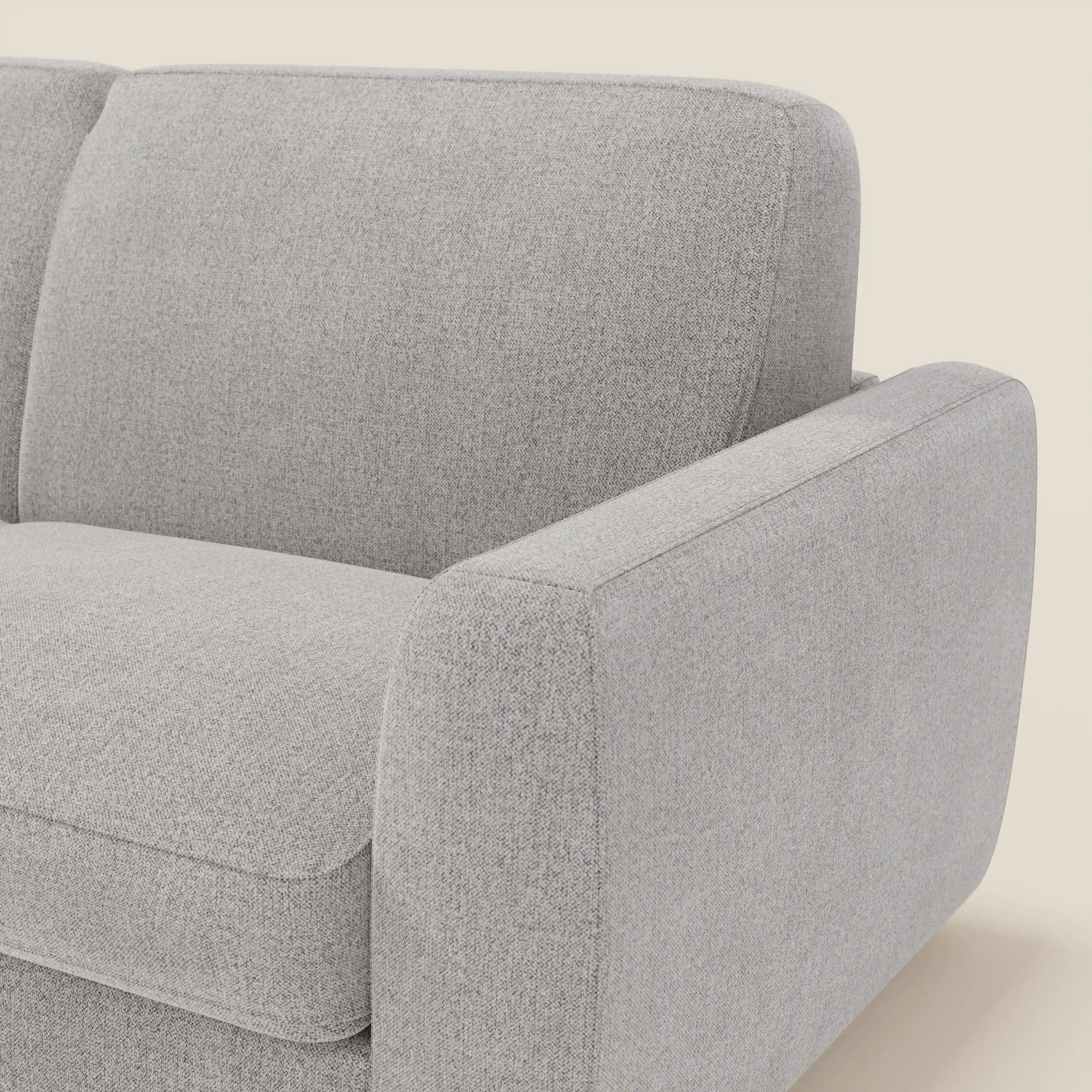 Un primo piano del divano letto sfoderabile Leonida di Divani.Store in tessuto Smacchiabile T23 grigio mostra lo schienale, il cuscino di seduta e il bracciolo destro. La morbida imbottitura ha una finitura liscia e leggermente strutturata. Sfondo: bianco sporco tinta unita.