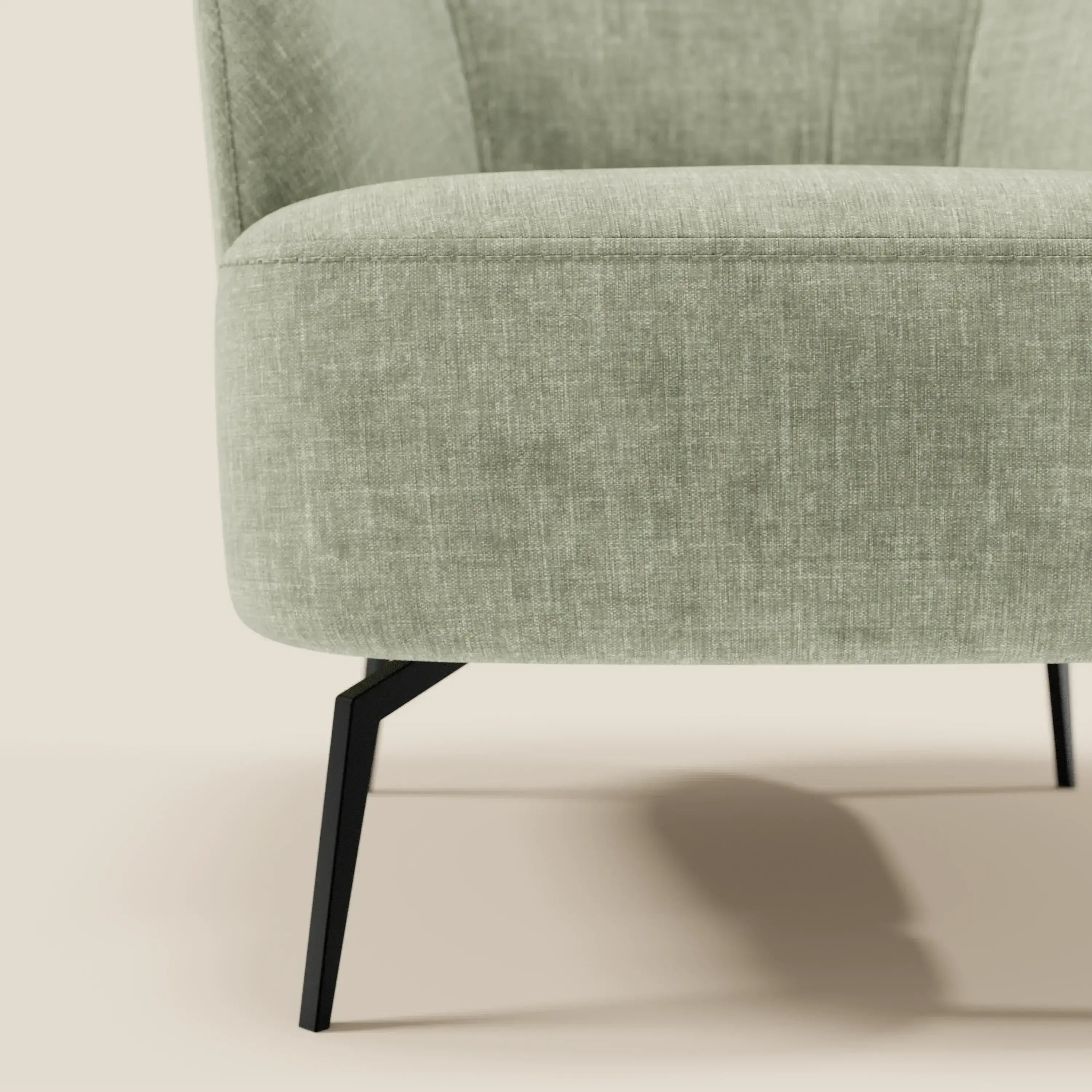 Primo piano della poltroncina Renoa di Divani.Store, un'elegante poltrona moderna in tessuto antimacchia verde salvia T24 con seduta arrotondata e gambe in metallo nero. In parte, è presentata su uno sfondo beige chiaro per un look minimalista e raffinato.