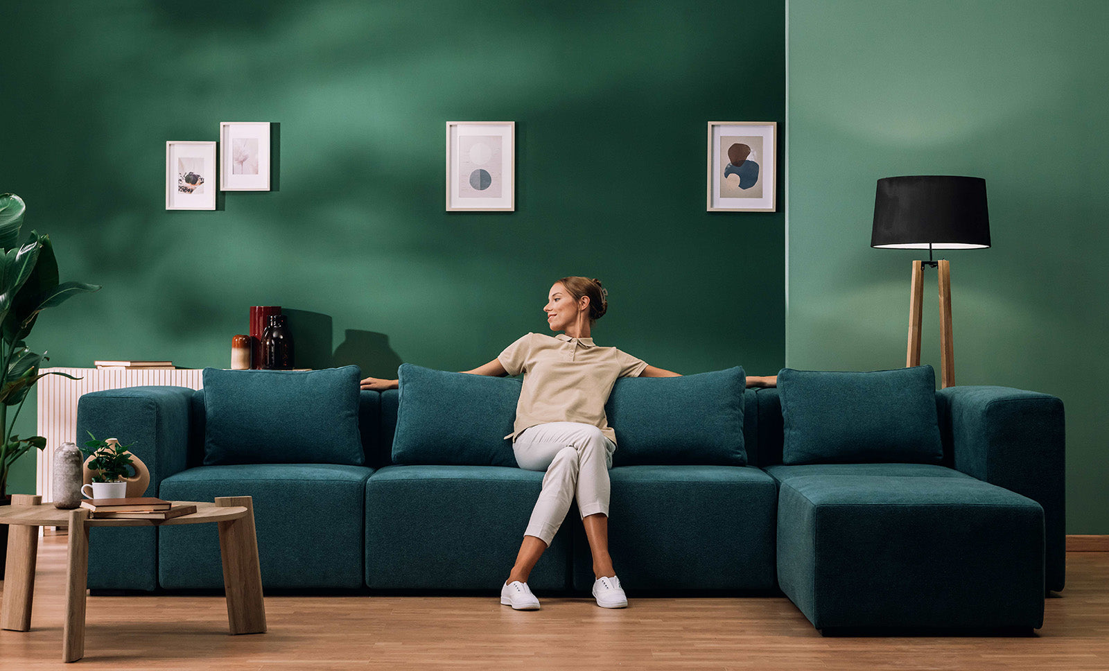 Donna seduta su un divano componibile verde in un soggiorno moderno con pareti verdi, pavimento in legno, opere d'arte astratte incorniciate, piante in vaso e lampada da terra. Arredamento accogliente e minimalista.