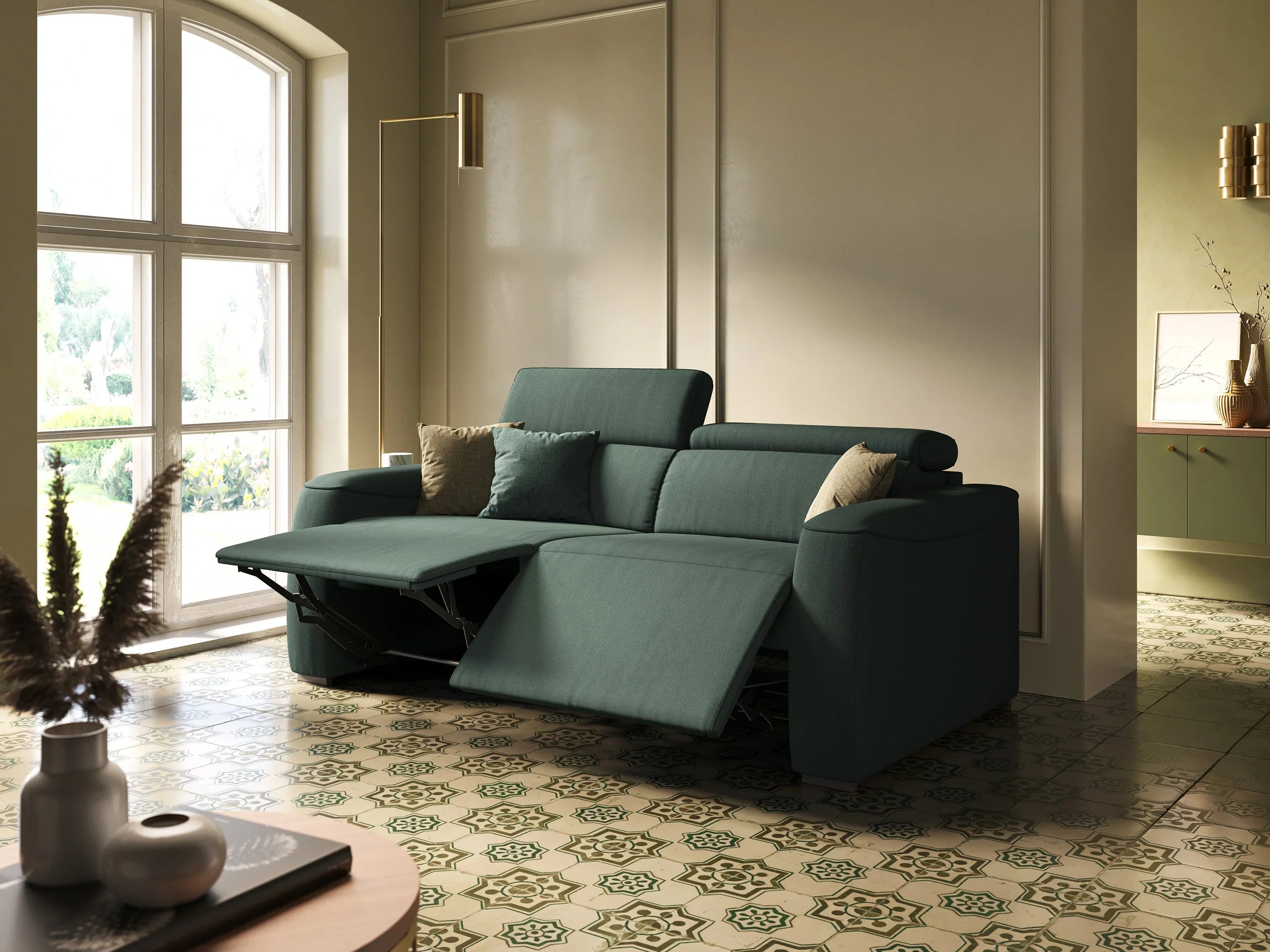 Un divano componibile verde acqua con poggiapiedi reclinati si trova su un pavimento di piastrelle a motivi vicino a una grande finestra, da cui entra la luce del sole. Accanto ci sono cuscini beige e verdi e una lampada da terra.