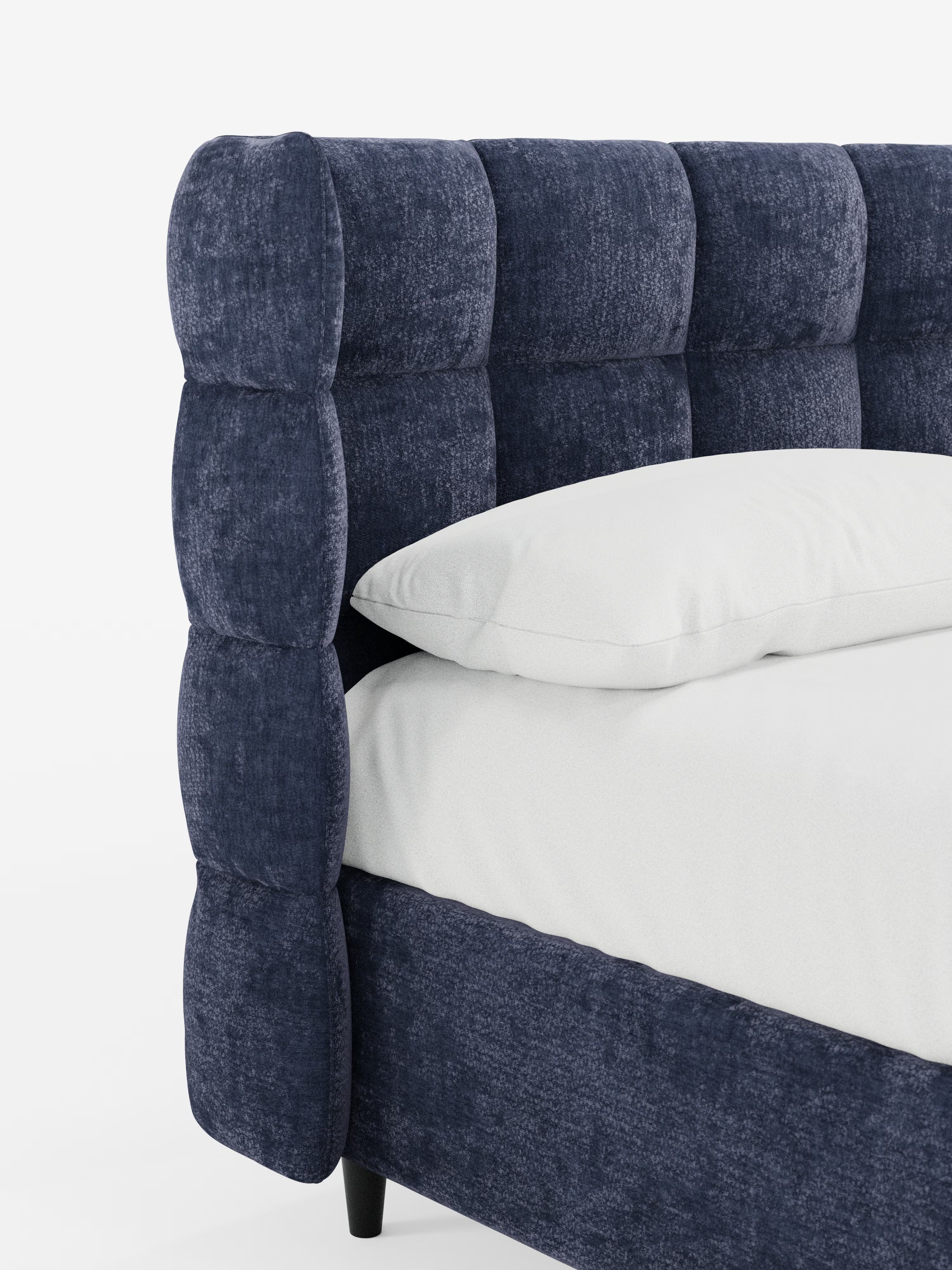 Un primo piano del letto imbottito RAGNAR di Letti.store mette in risalto il suo elegante tessuto blu navy certificato Oeko-Tex® e una testiera capitonné avvolgente, spessa e trapuntata, con pannelli laterali abbinati. Spicca il rivestimento morbido e dalla trama ricca, mentre sul materasso poggiano un cuscino bianco e un lenzuolo. Lo sfondo è bianco a tinta unita.