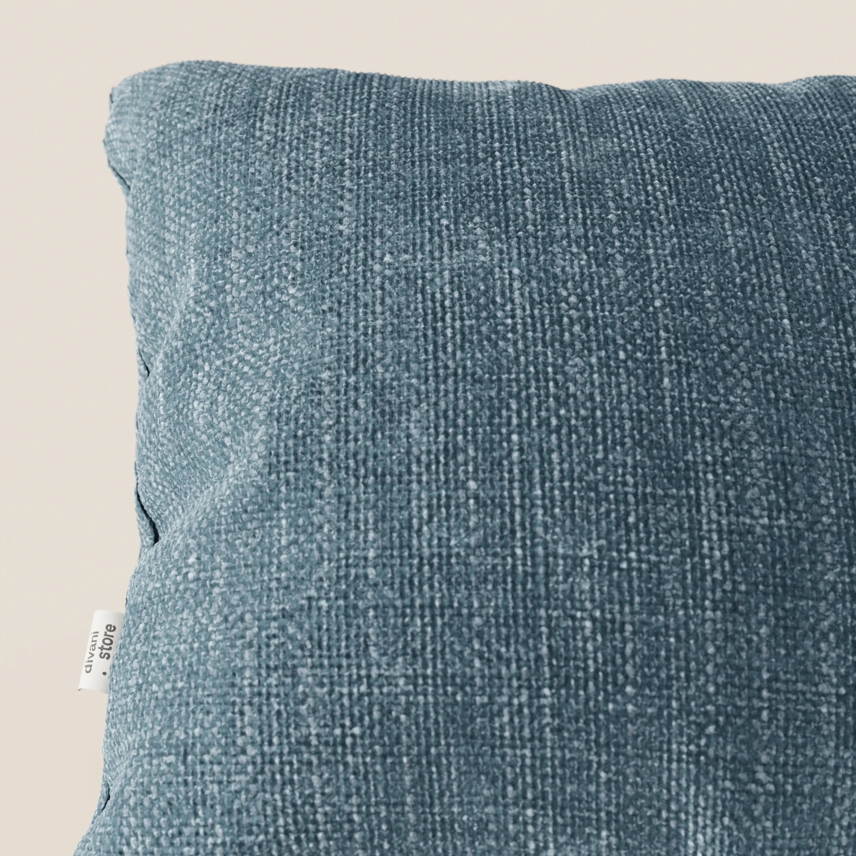 Primo piano del cuscino Lyon 45x45 cm in T06 azzurro di Divani.Store, realizzato in morbido tessuto intrecciato impermeabile, presentato su uno sfondo beige con una piccola etichetta bianca con testo nero sul lato sinistro.