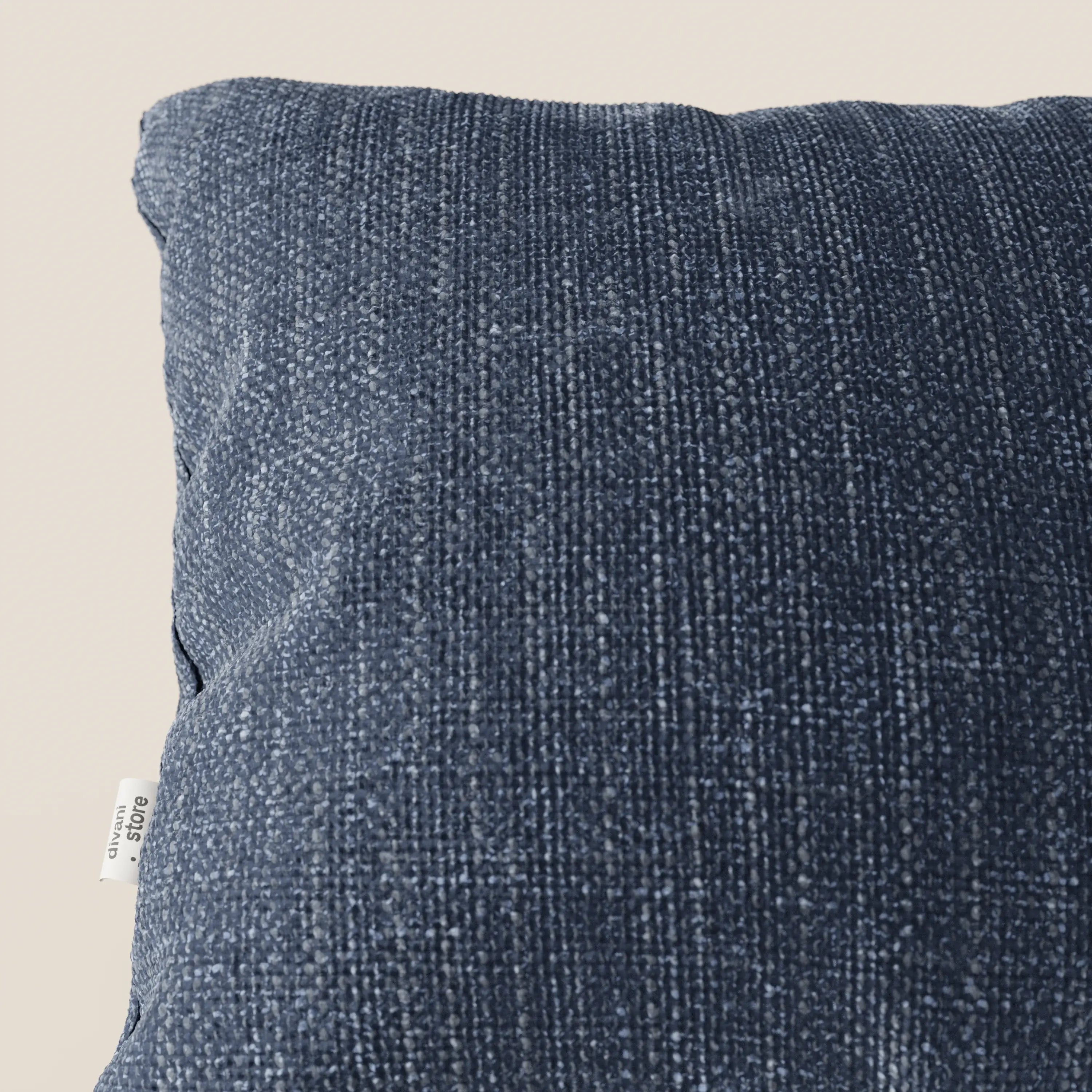 Primo piano del cuscino Lyon di Divani.Store in T06 blu, realizzato in morbido tessuto impermeabile. Su uno sfondo beige chiaro, sul bordo sinistro compare una piccola etichetta bianca. Il cuscino sembra soffice ed è leggermente inclinato verso la macchina fotografica.