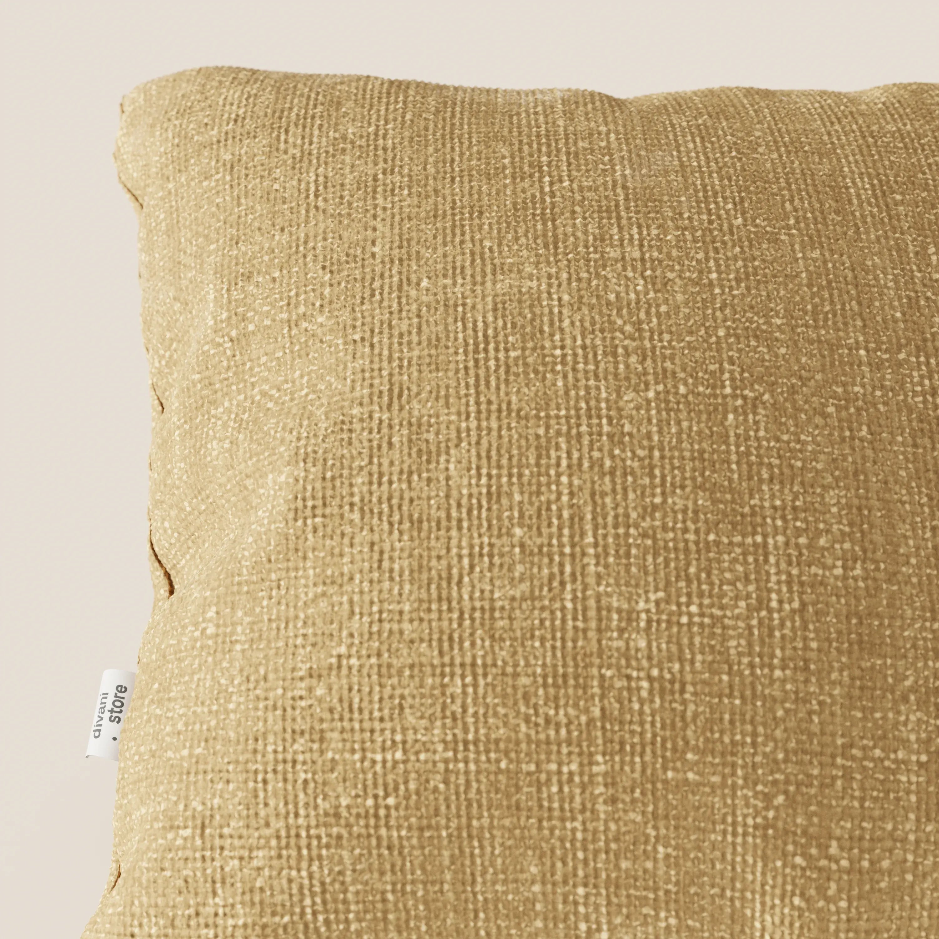 Primo piano del cuscino Lyon di Divani.Store in ocra T06, realizzato in morbido tessuto intrecciato impermeabile. La superficie gialla strutturata spicca su uno sfondo beige; sulla cucitura sinistra è visibile un'etichetta bianca con testo nero.