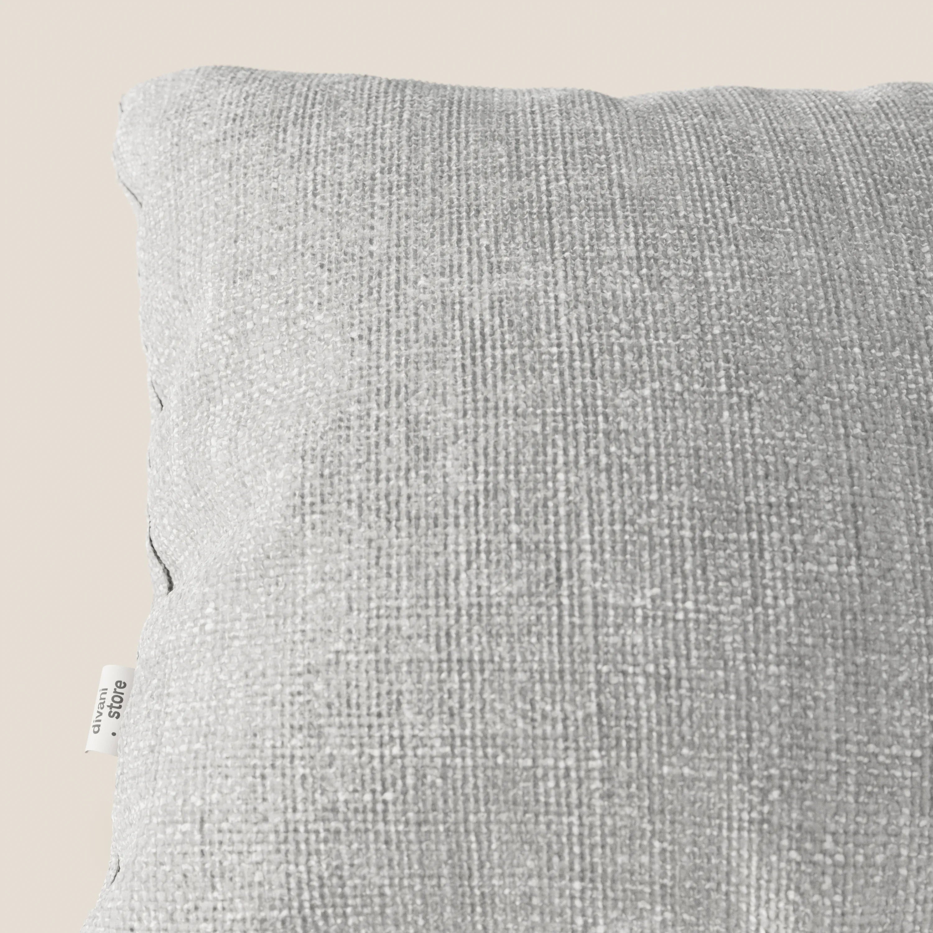 Primo piano del cuscino Lyon 45x45 cm di Divani.Store in tessuto impermeabile intrecciato T06 grigio, che mostra la trama a vista e l'etichetta laterale bianca sul bordo sinistro su sfondo beige. È visibile solo una parte del cuscino.
