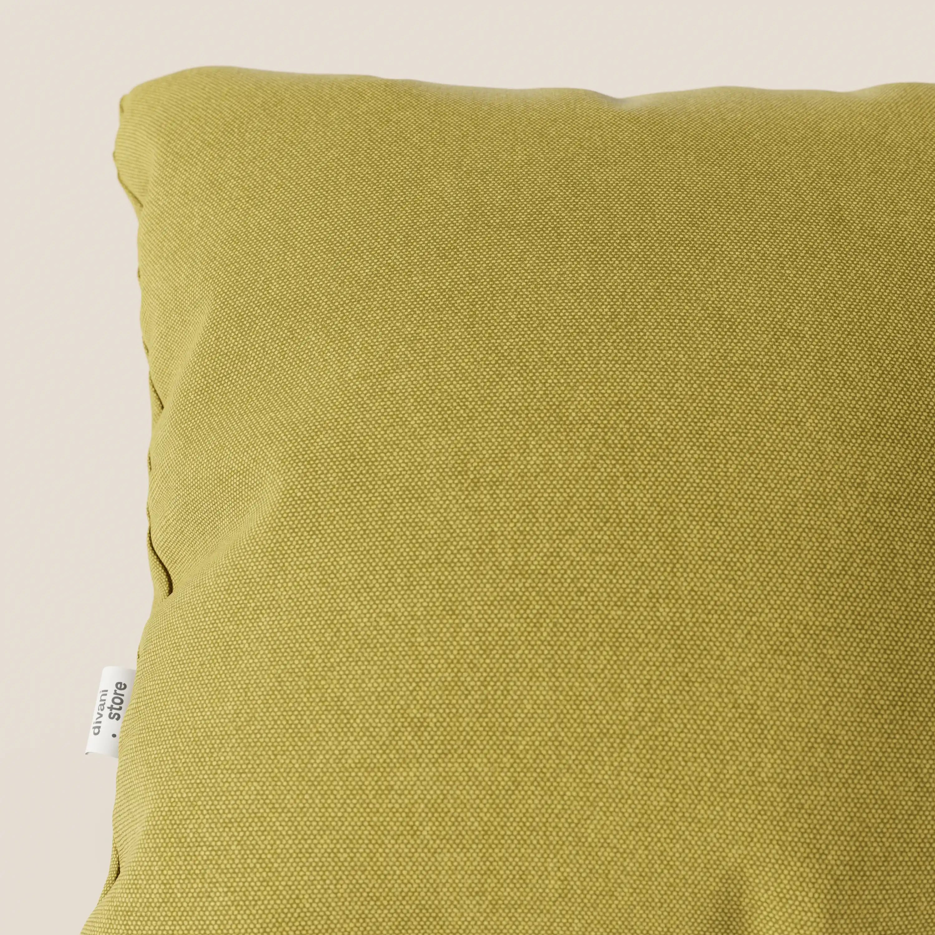 Primo piano del cuscino Divani.Store Honey in giallo T13, realizzato in tessuto simil-cotone resistente all'acqua, con una piccola etichetta laterale bianca. Il cuscino quadrato è inserito in uno sfondo beige chiaro.