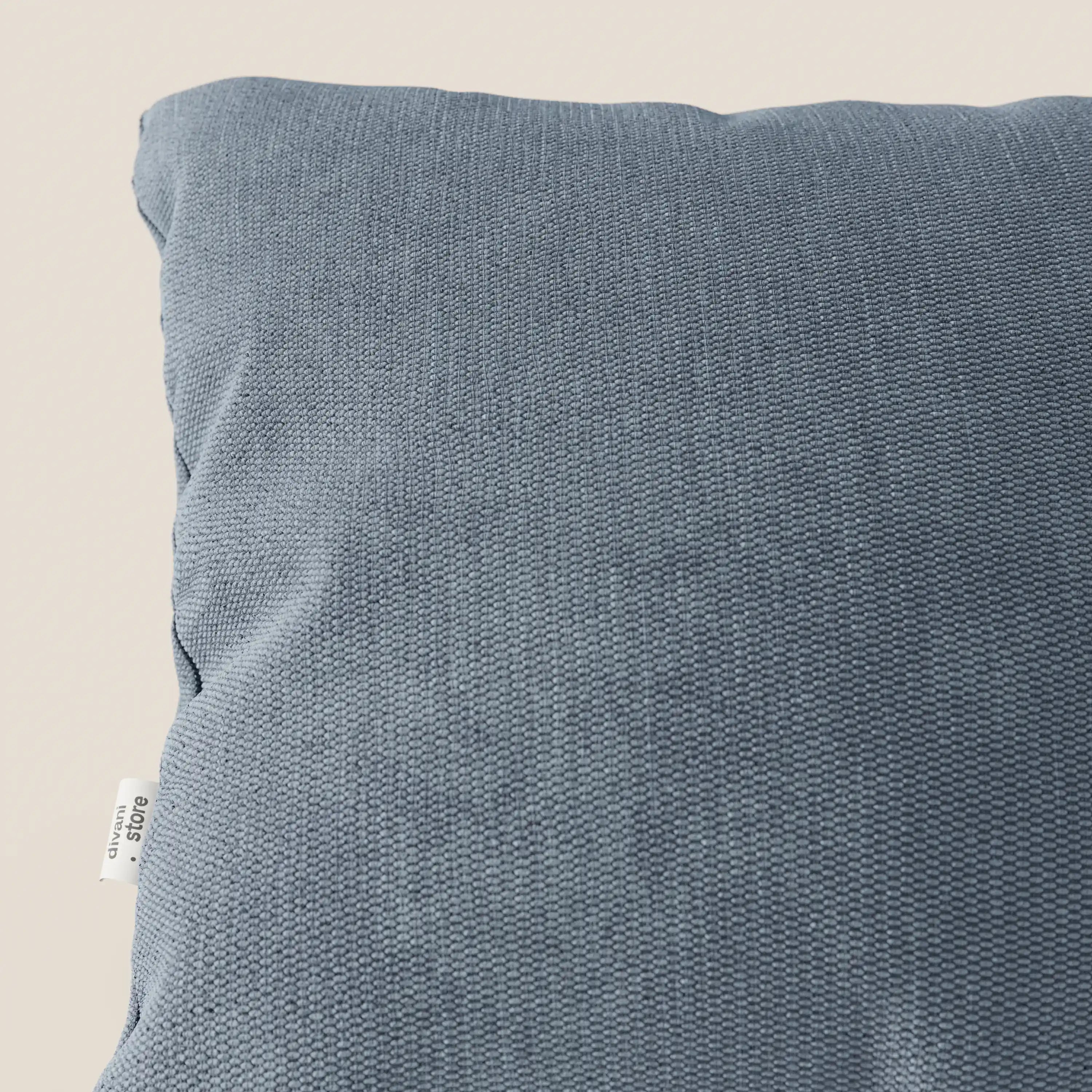Primo piano dell'angolo superiore sinistro del cuscino Miami di Divani.Store in tessuto impermeabile T14 carta da zucchero, con un piccolo cartellino bianco di manutenzione, ideale per cuscini decorativi con fodera lavabile e facili da pulire.