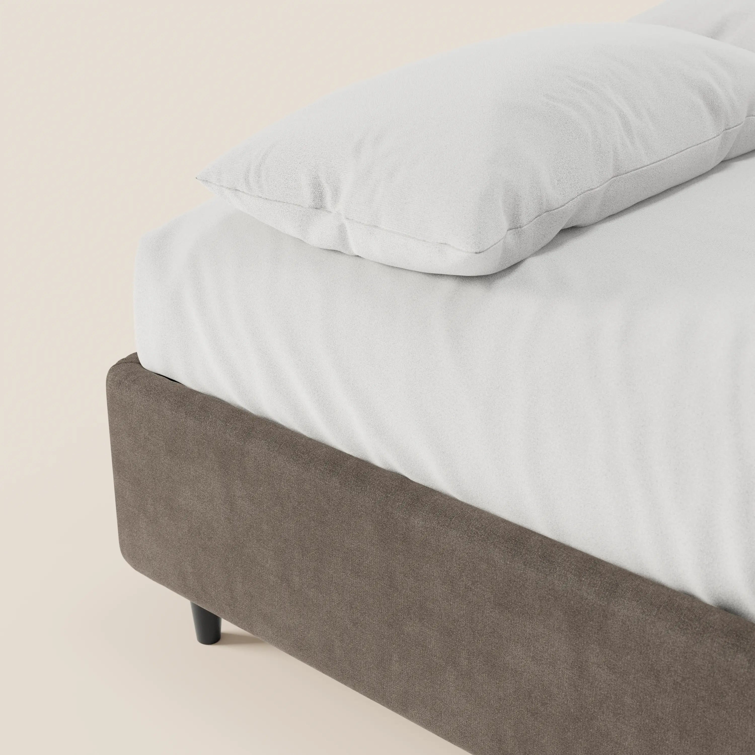 Un primo piano del letto sommier imbottito ERON di Letti.store, con lenzuolo e cuscino bianchi su una struttura rivestita in tessuto marrone simile alla pelle scamosciata, in tessuto impermeabile certificato Oeko-Tex, con gambe corte nere e senza testiera, su uno sfondo color crema chiaro.
