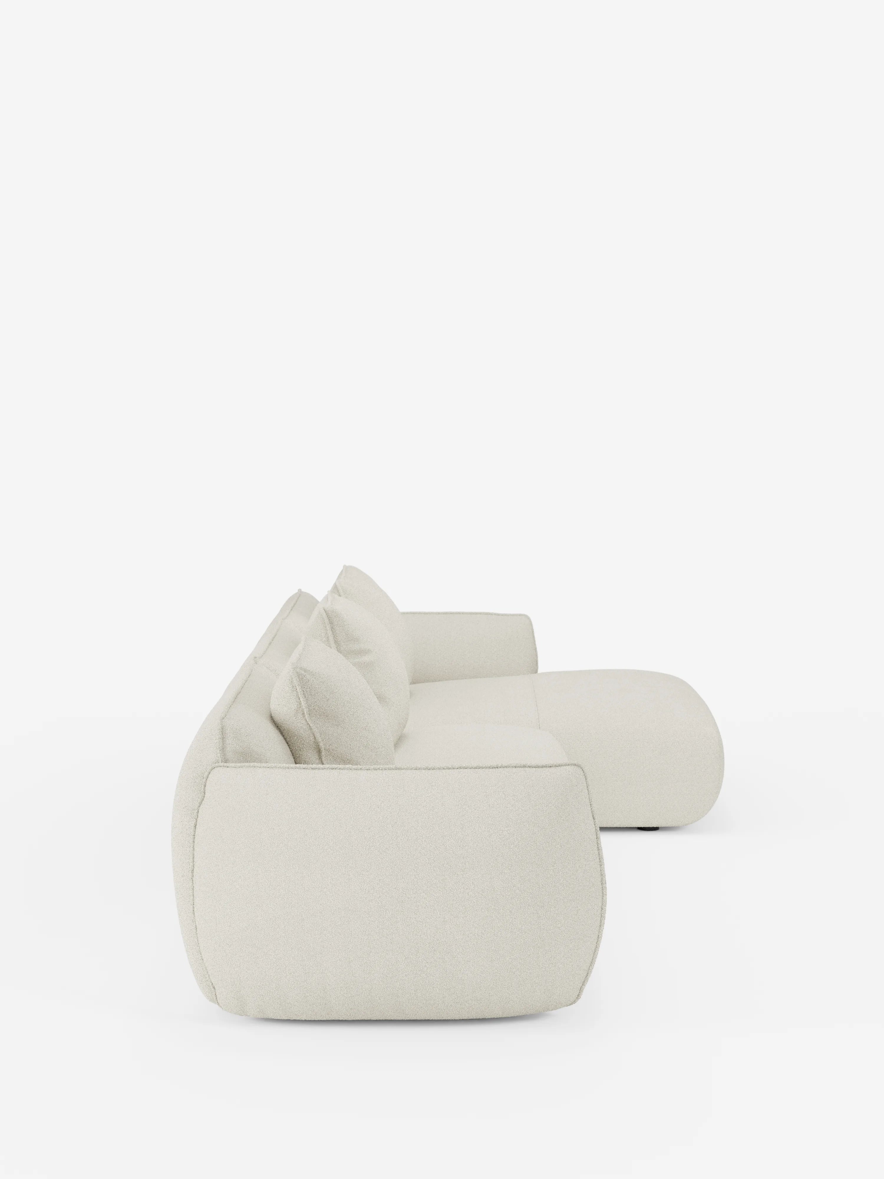 Vista laterale del divano angolare con penisola componibile Nimbus in morbido tessuto effetto cotone smacchiabile T33 di Divani.Store, un divano moderno e minimalista color bianco sporco con bordi arrotondati e morbidi cuscini che risaltano su uno sfondo bianco a tinta unita. Questo modello contemporaneo presenta un profilo basso, due cuscini dello schienale e un ampio bracciolo per un maggiore comfort.