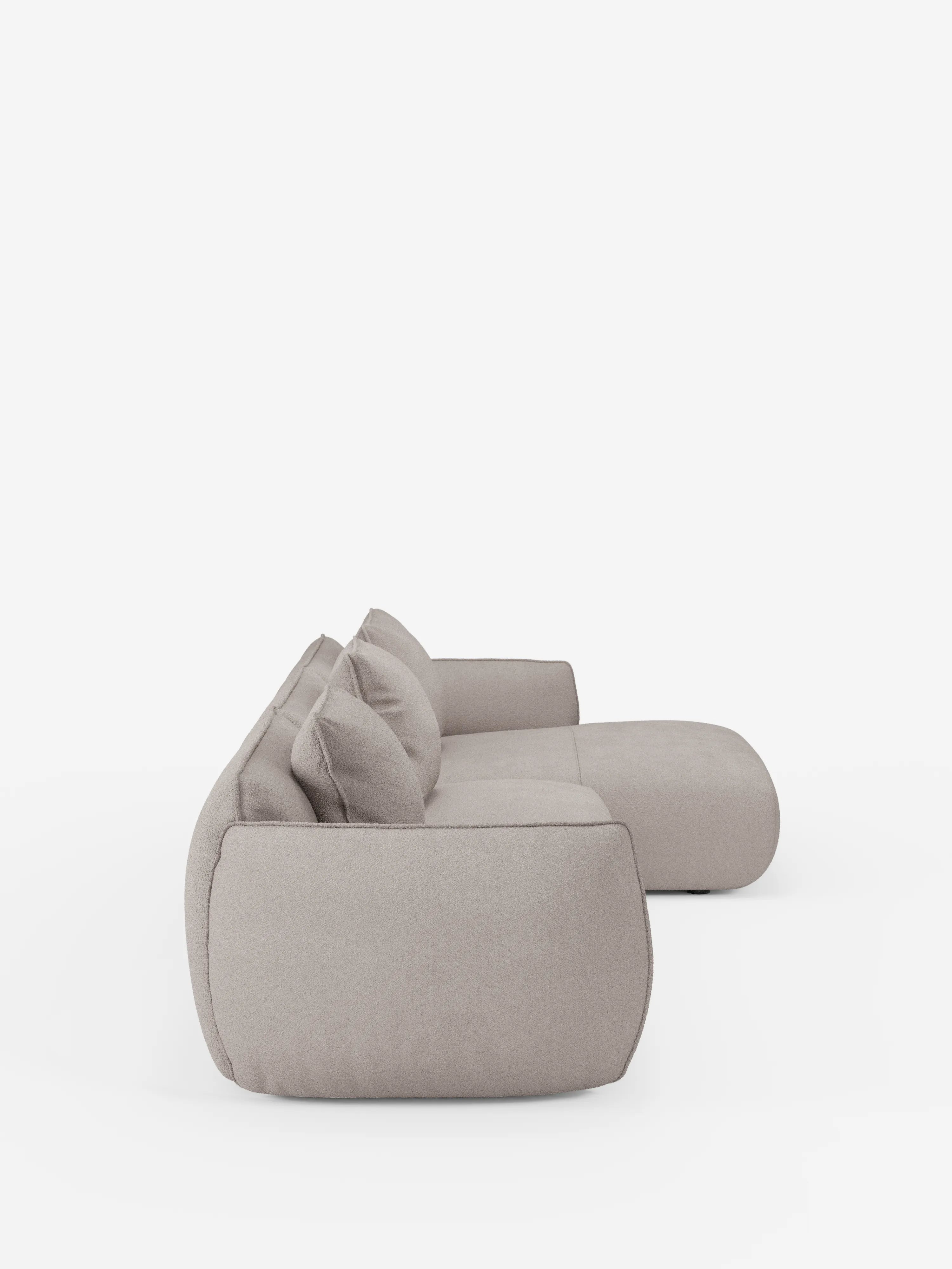 Il divano angolare con penisola componibile Nimbus in morbido tessuto effetto cotone smacchiabile T33 di Divani.Store è un moderno divano componibile color beige chiaro, ripreso in una vista laterale, caratterizzato da cuscini spessi e arrotondati e da due cuscini dello schienale abbinati. Sullo sfondo bianco a tinta unita, la sua texture morbida e imbottita e il suo design contemporaneo e delicato risaltano ancora di più.