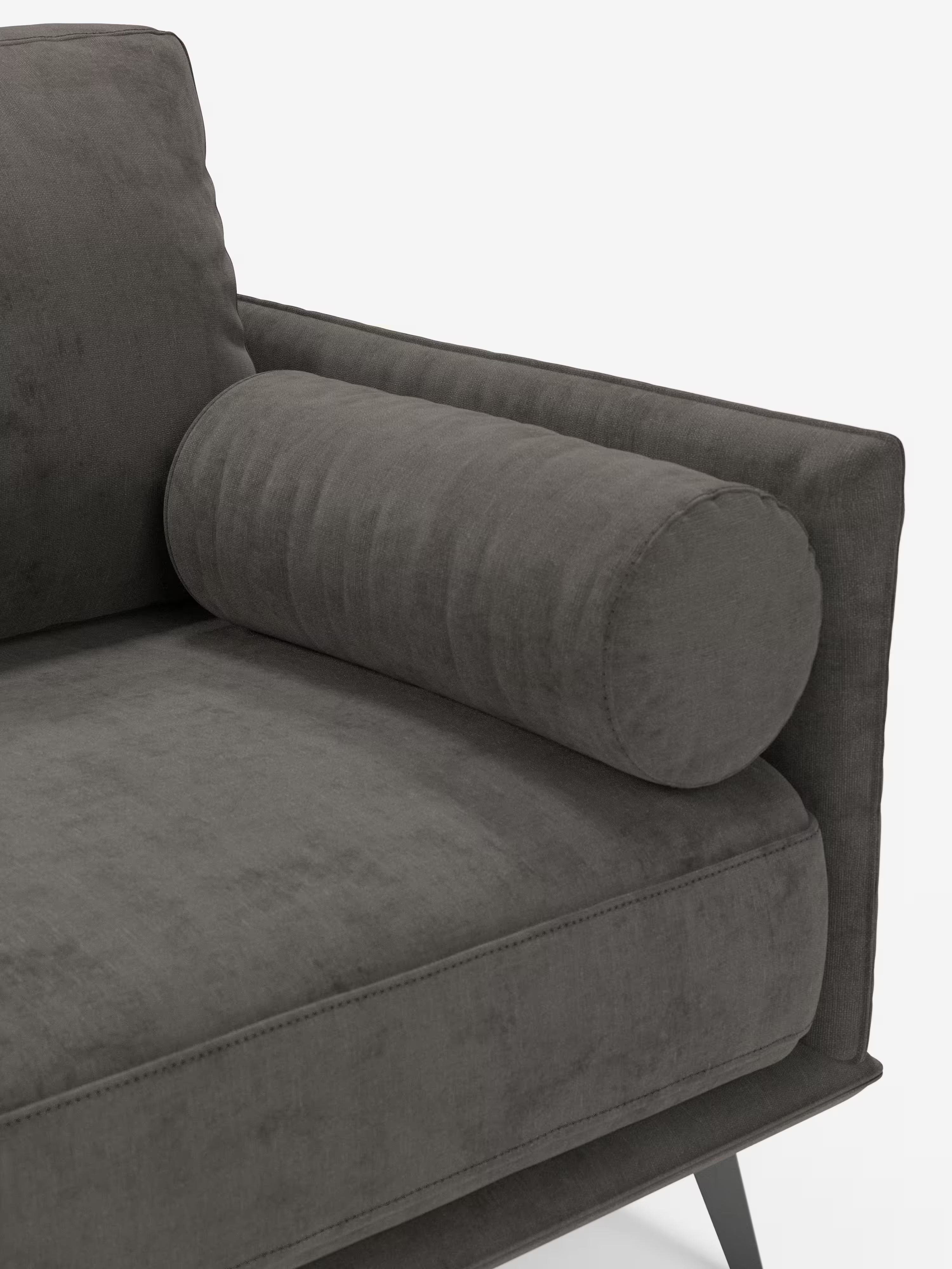 Primo piano del divano lineare a 2 posti "Basalto" di Divani.Store in grigio scuro, che mette in risalto parte della seduta, dello schienale e dei braccioli senza cuciture, con un cuscino cilindrico coordinato. Rivestito in morbido tessuto T31 resistente alle macchie e dotato di un pratico rivestimento sfoderabile che ne facilita la manutenzione, questo divano moderno presenta eleganti gambe in metallo angolate, perfette per un’estetica minimalista. Sfondo bianco.