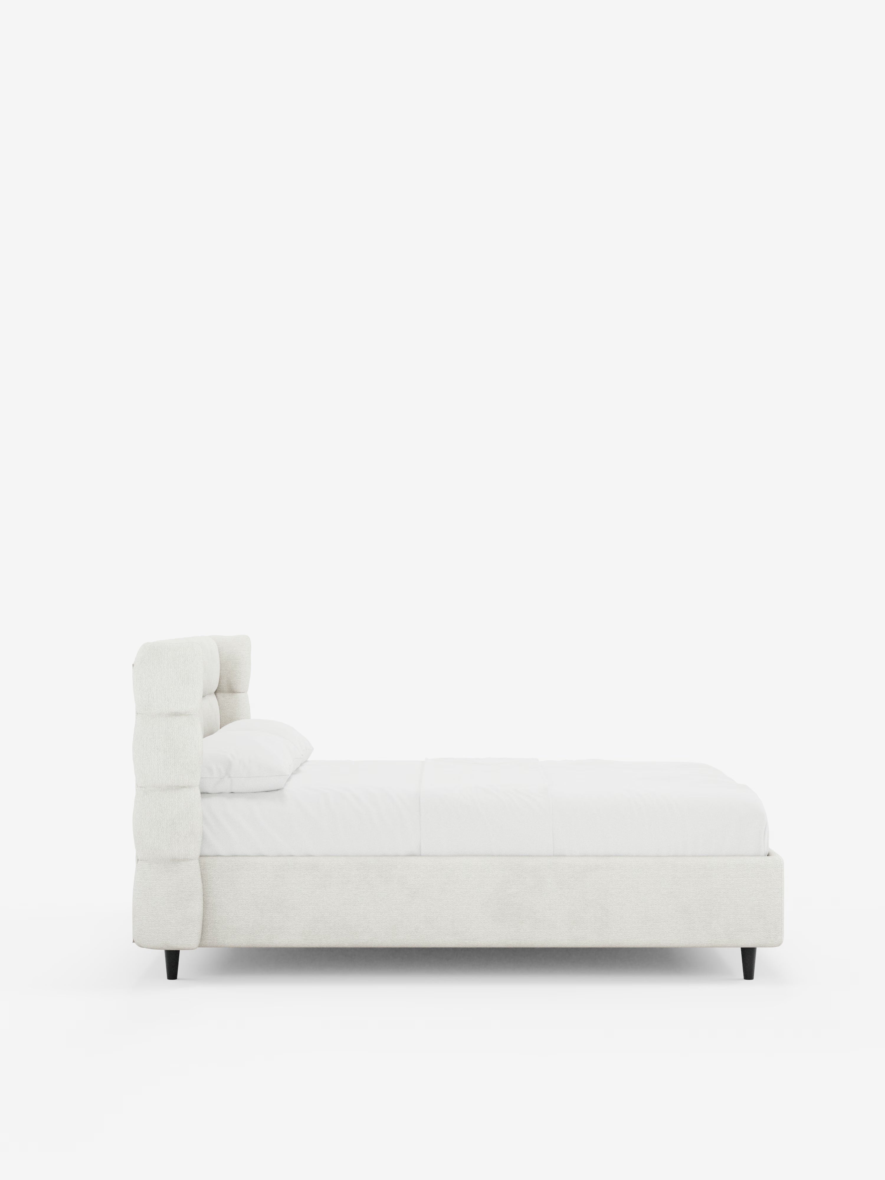 Vista laterale del letto imbottito RAGNAR con contenitore e testiera capitonné avvolgente di Letti.store, caratterizzato da un’alta testiera imbottita avvolgente realizzata in un elegante tessuto bianco sporco certificato Oeko-Tex®. Il letto è abbinato a biancheria da letto bianca e a un unico cuscino, ed è sostenuto da gambe corte e scure. È posizionato su uno sfondo bianco a tinta unita per enfatizzarne il design pulito e minimalista.