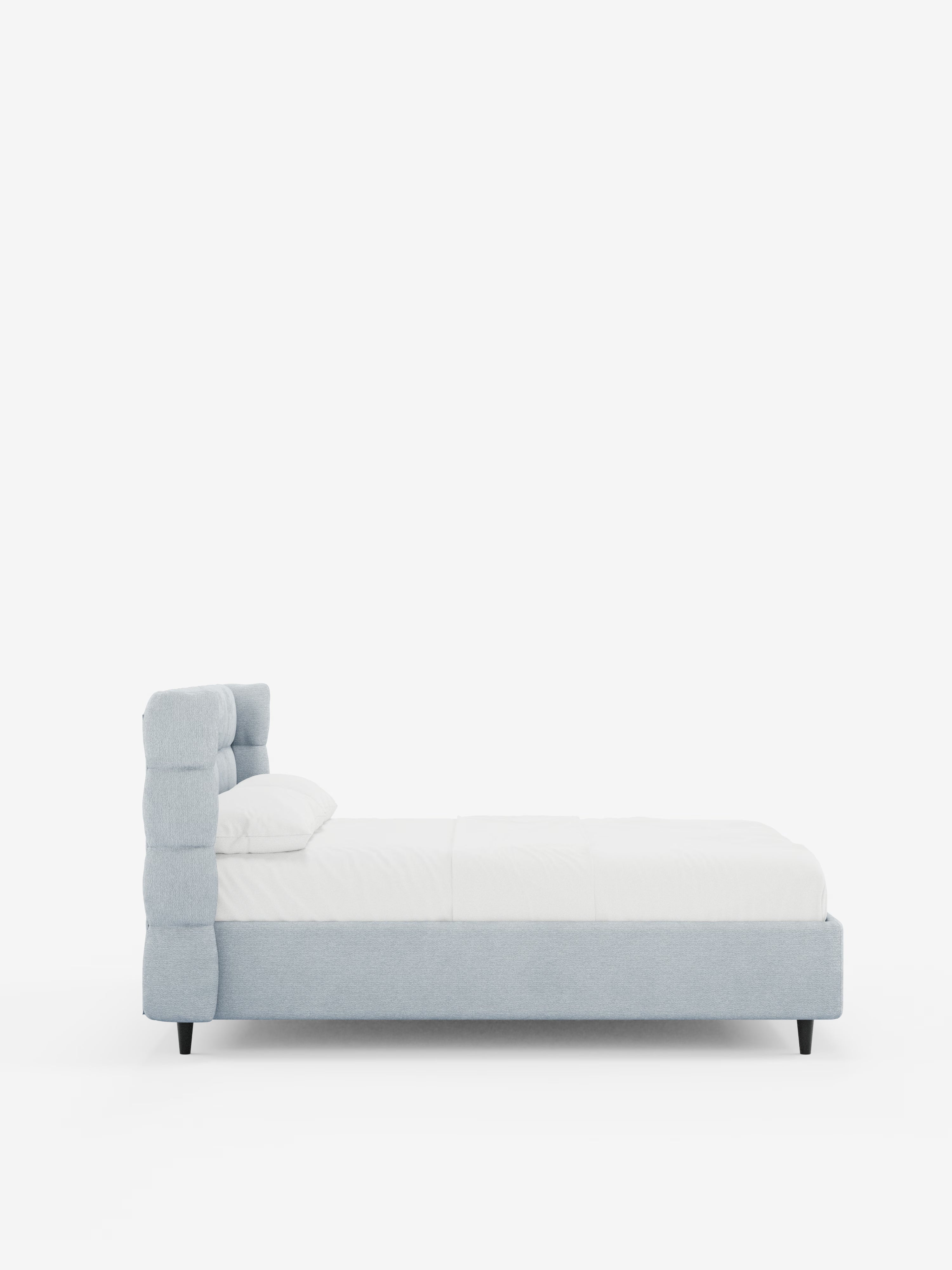 Vista laterale del letto imbottito RAGNAR con contenitore e testiera capitonné avvolgente di Letti.store, rivestito in un elegante tessuto azzurro certificato Oeko-Tex®. Il letto è abbinato a biancheria da letto bianca e a un unico cuscino, mentre sotto la struttura sono visibili le gambe corte di colore nero. Uno sfondo bianco a tinta unita ne esalta il design pulito e minimalista.