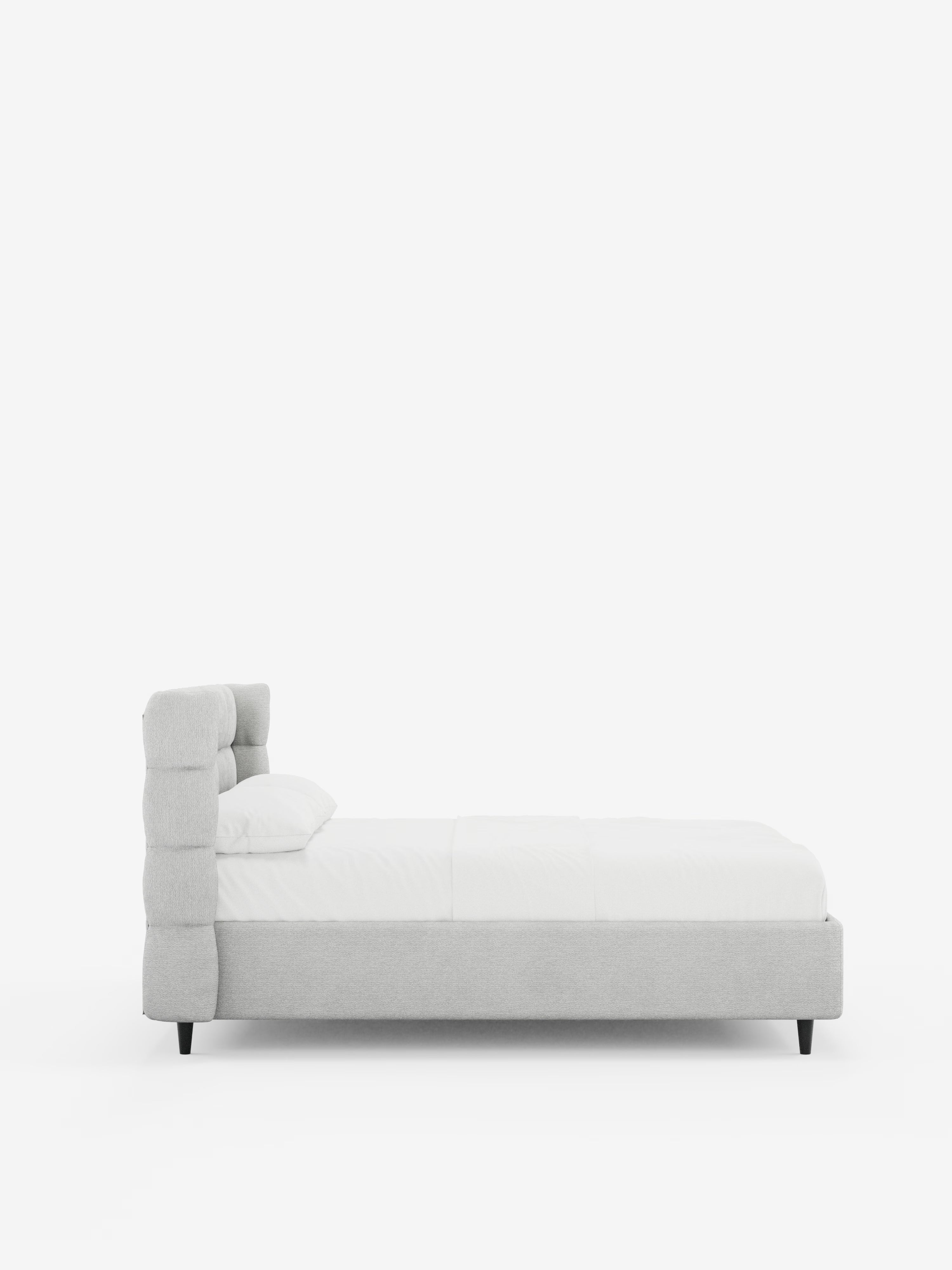 Vista laterale del letto imbottito RAGNAR con contenitore e testiera capitonné avvolgente di Letti.store, caratterizzato da una testiera capitonné avvolgente di colore grigio chiaro, realizzata in elegante tessuto certificato Oeko-Tex®, biancheria da letto bianca e gambe nere su uno sfondo bianco a tinta unita. Il cuscino e il materasso sono disposti con cura, mettendo in risalto il design pulito e minimalista del letto.